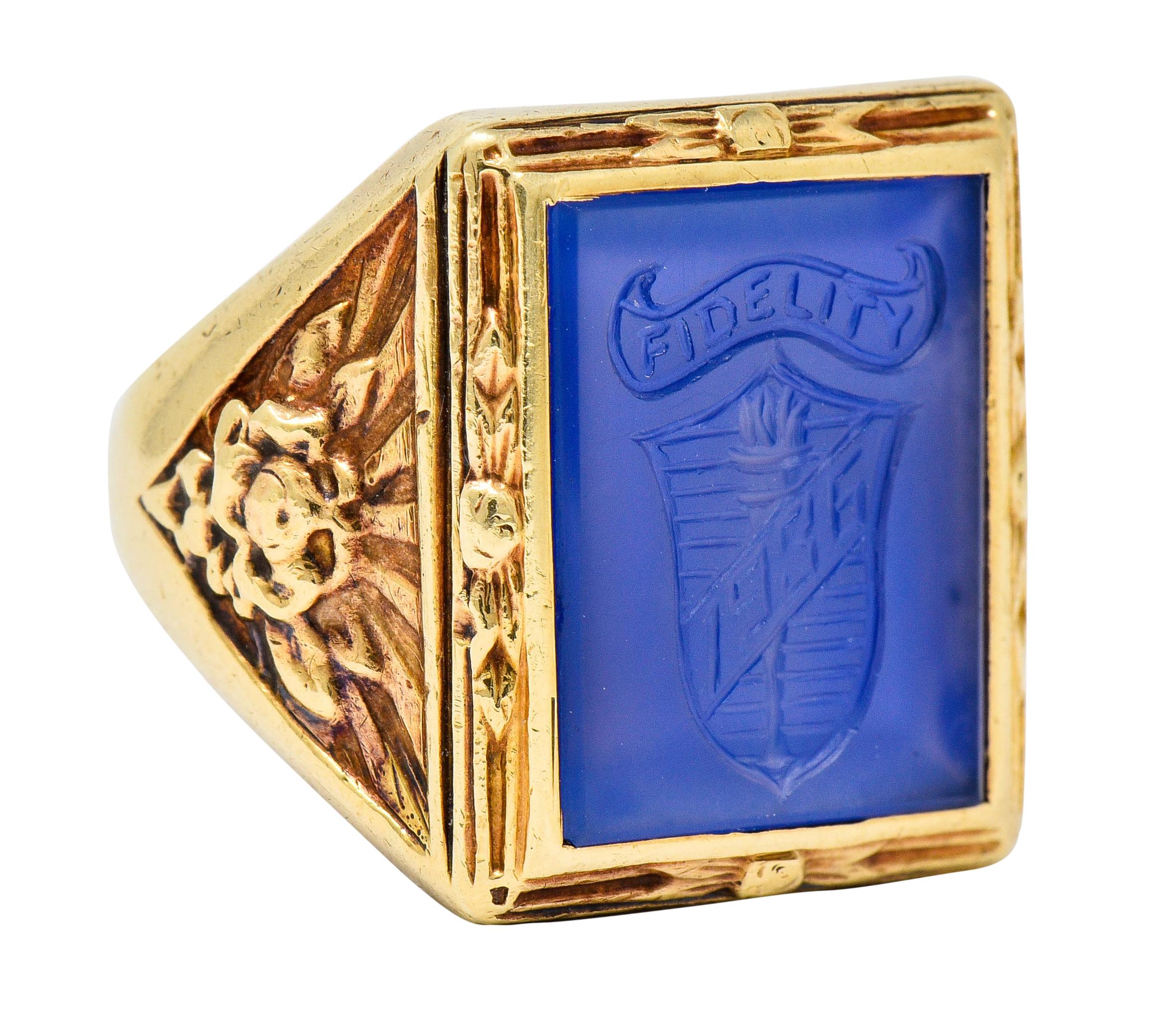 Centering a bezel set rectangular intaglio tablet of chalcedony measuring approximately 14.0 x 11.0 mm

Bright blue in color, dyed, and deeply engraved to depict a striped shield emblazoned by a torch and dated '1936'

Topped by a scrolled ribbon