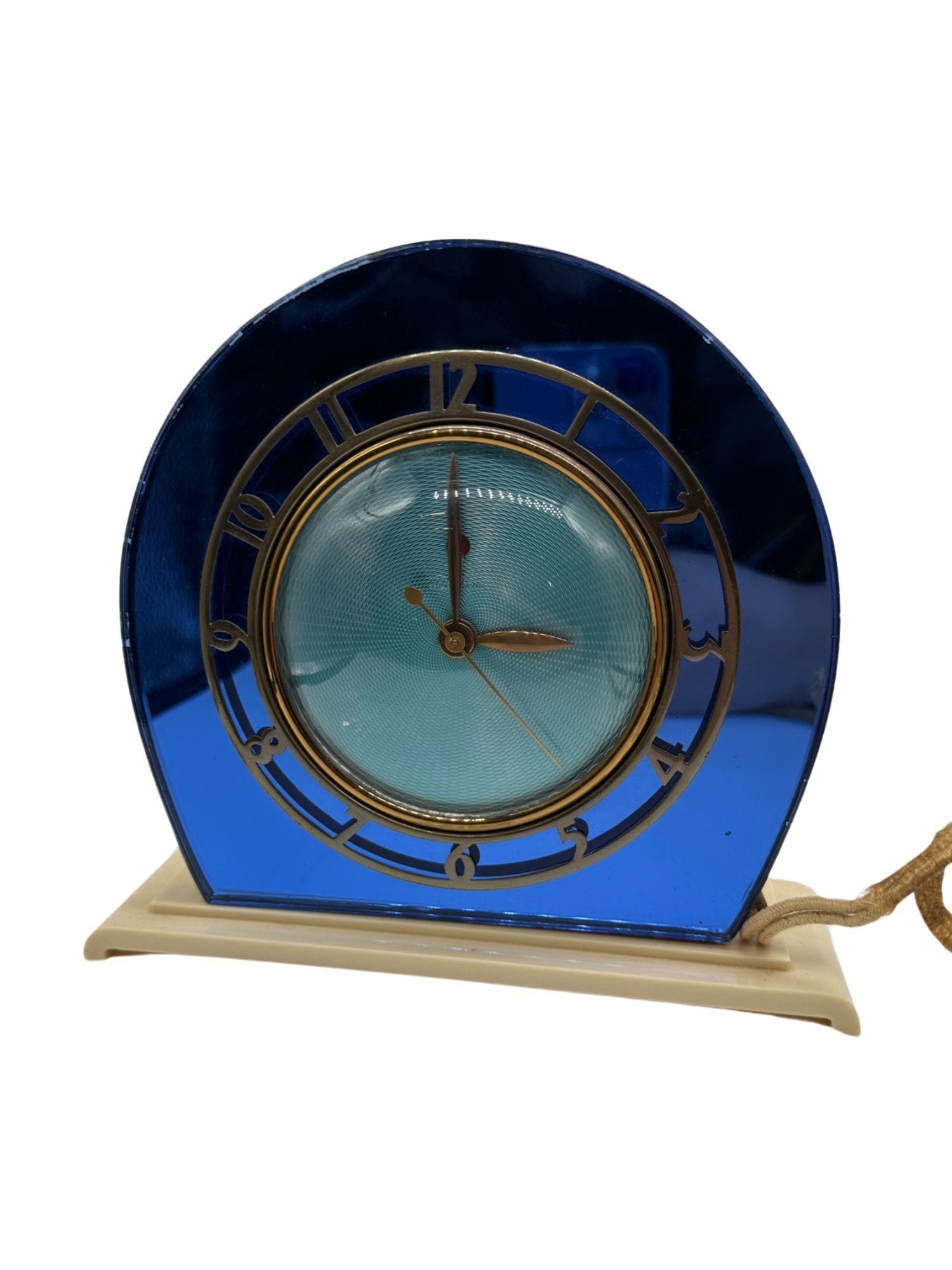 It features a stunning cobalt blue mirror, complemented by a polished white Plaskon base and hands. The dial boasts a textured blue surface, while the polished brass bezel is adorned with cut-out numerals. To adjust the time, simply pull out and