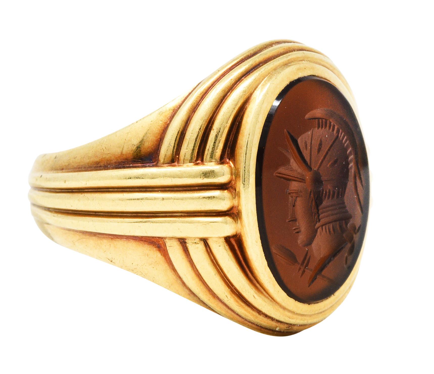 Signet style ring centers an oval intaglio of sard carnelian - measures 15.0 x 11.0 mm

Medium dark brownish red and deeply engraved to depict the profile of a Greek warrior

Bezel set in a deeply grooved mounting

Stamped 14 for 14 karat