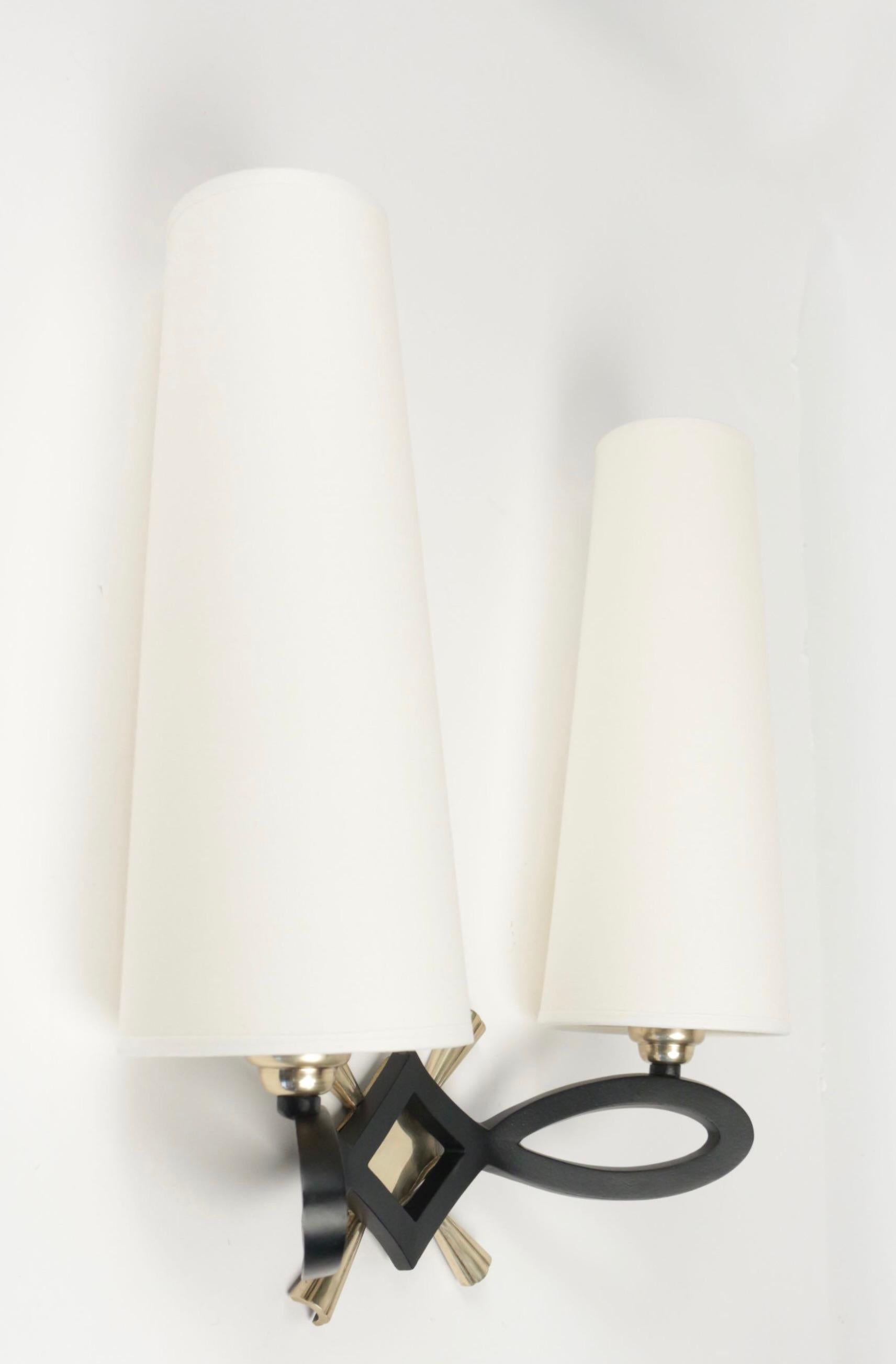 Each sconce is composed of two loops in black lacquered bronze underlined by a plate and a diamond-shaped cross in gilded brass serving as a wall support. 
They are dressed with two lampshades in off-white cotton redone identically distributed on