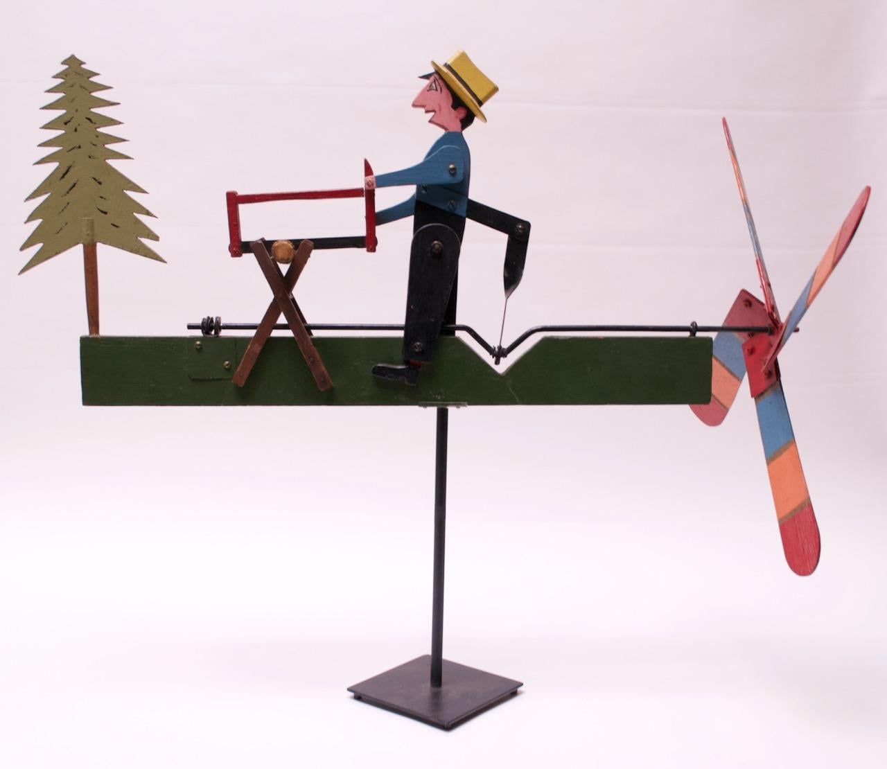 Whimsical whirligig depicting a woodsman sawing a log mounted to a black painted metal stem / base. Hand carved, painted, and assembled with applied facial details. When manually turned, the propeller activates the woodman's 'sawing' motion. The