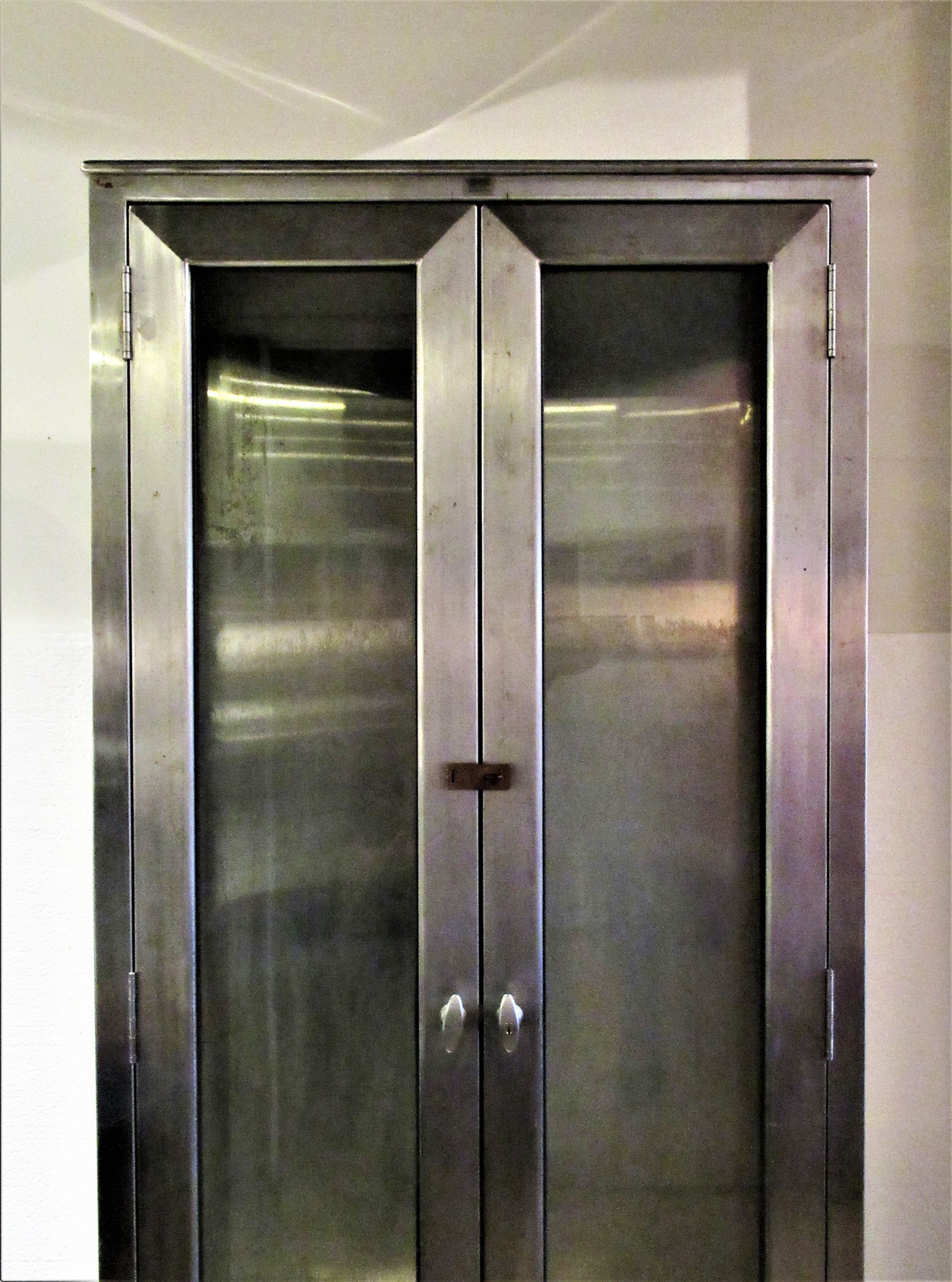 Tall sleek and narrow form stainless steel industrial medical cabinet having two locking glass front doors with panel glass sides and raised on four shaped angled legs, circa 1940s. Exceptional quality. This one's a beauty. See all pictures and read