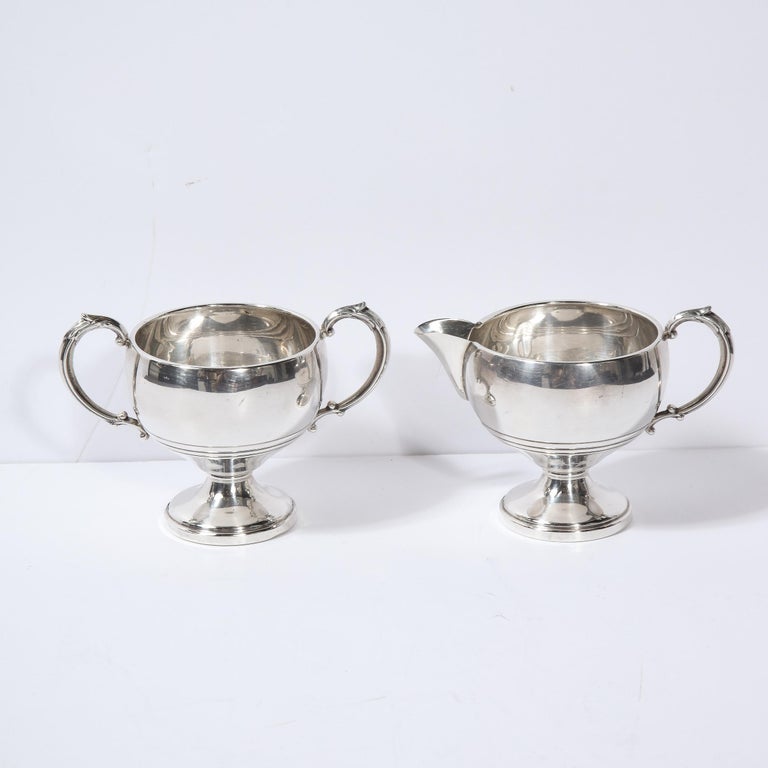 This elegant sugar and creamer set was realized in the United States circa 1940. It features a creamer with a circular base; a rounded body; and a streamlined handle. The sugar caddy is of a similar form with two streamlined handles on opposing