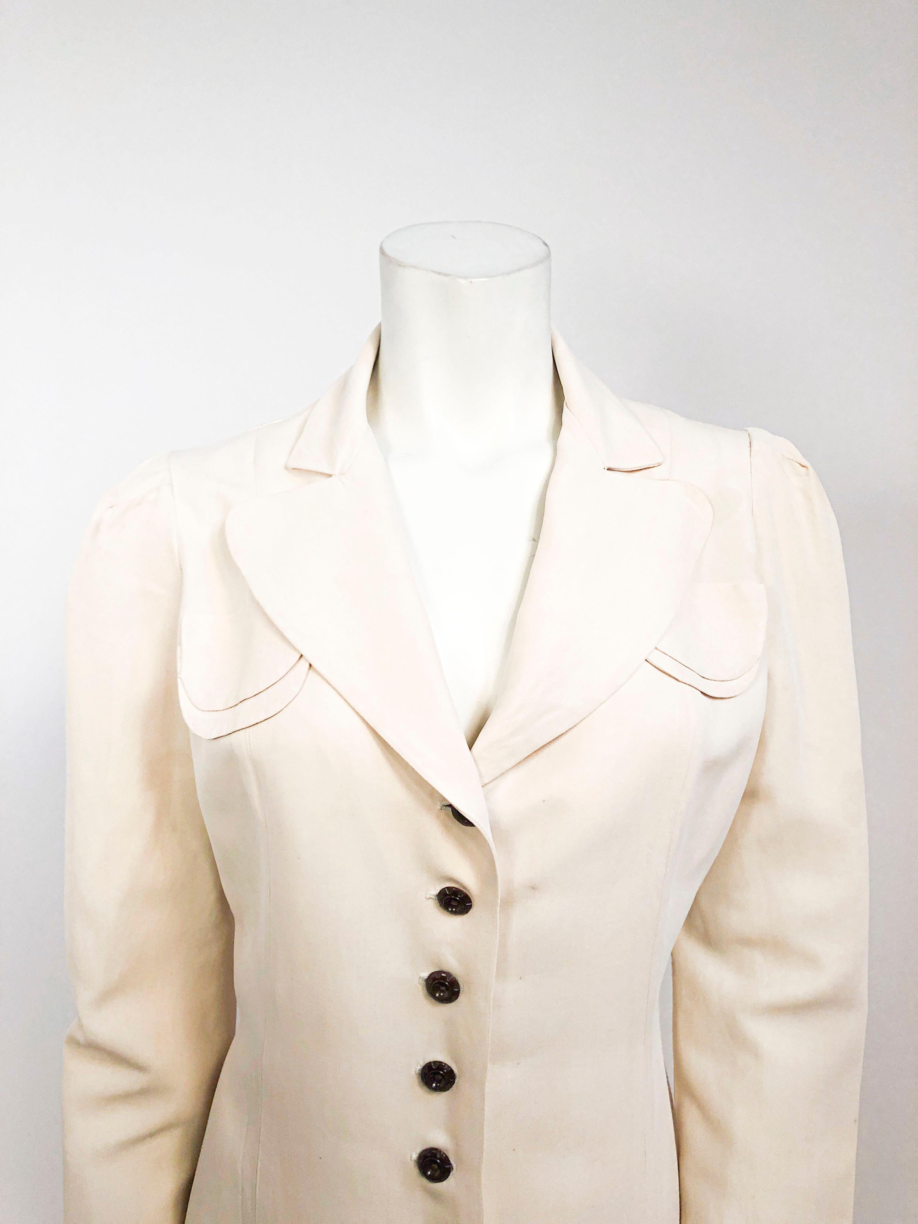 1940s coat in beige with a very light hue of pink. The textile is a fine rayon twill that is light-weight. This piece is decorated with faux pockets, enlarged collar,  and original brown buttons.The sleeves are gathered at the shoulder and darted at