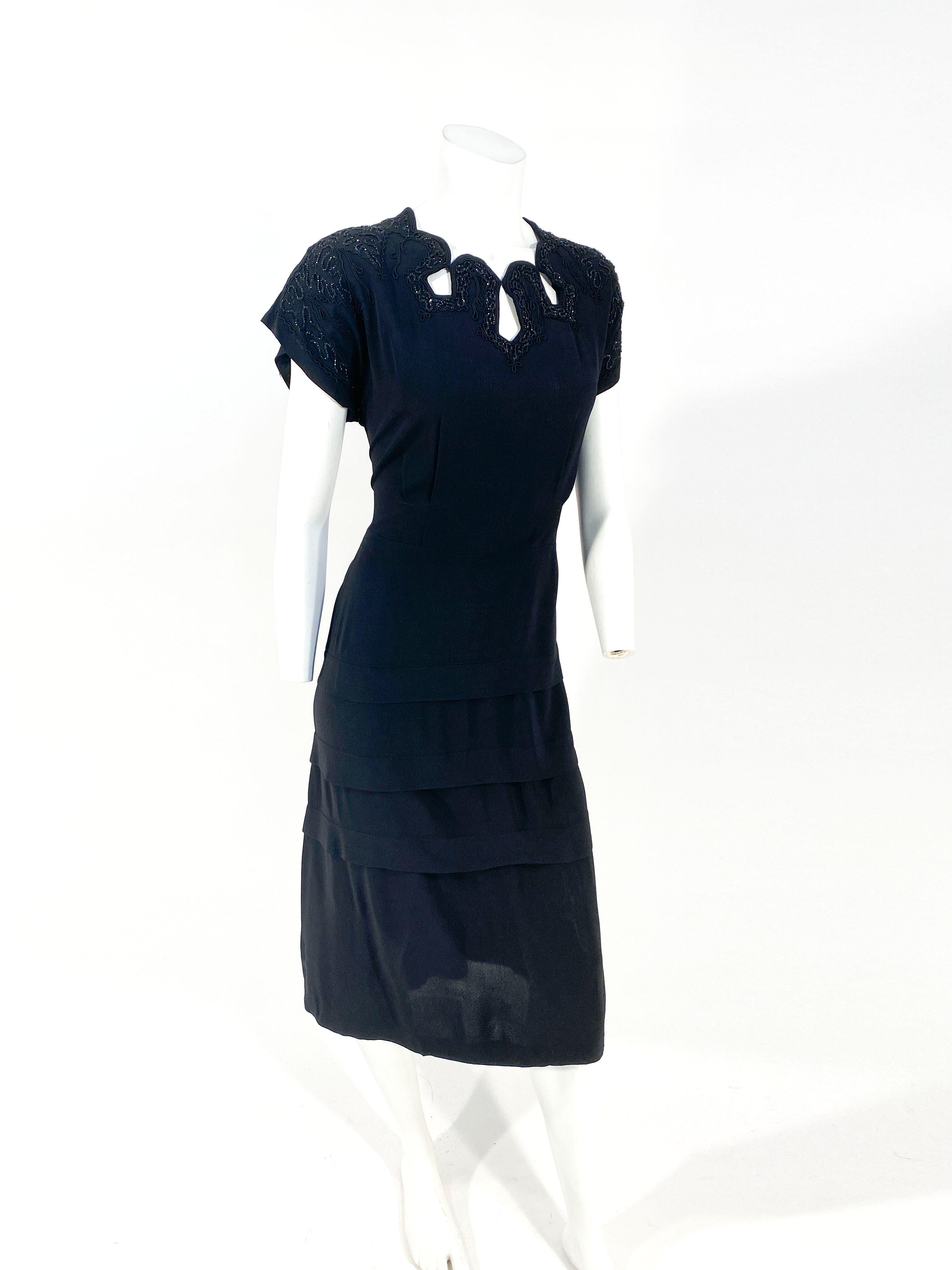 1940s black crepe cocktail dress featuring a decorative cutout neckline accented with soutache and beading extending the the cap sleeves. The skirt of the dress has three horizontal knife pleats. This dress has a side metal zipper and a back zipper