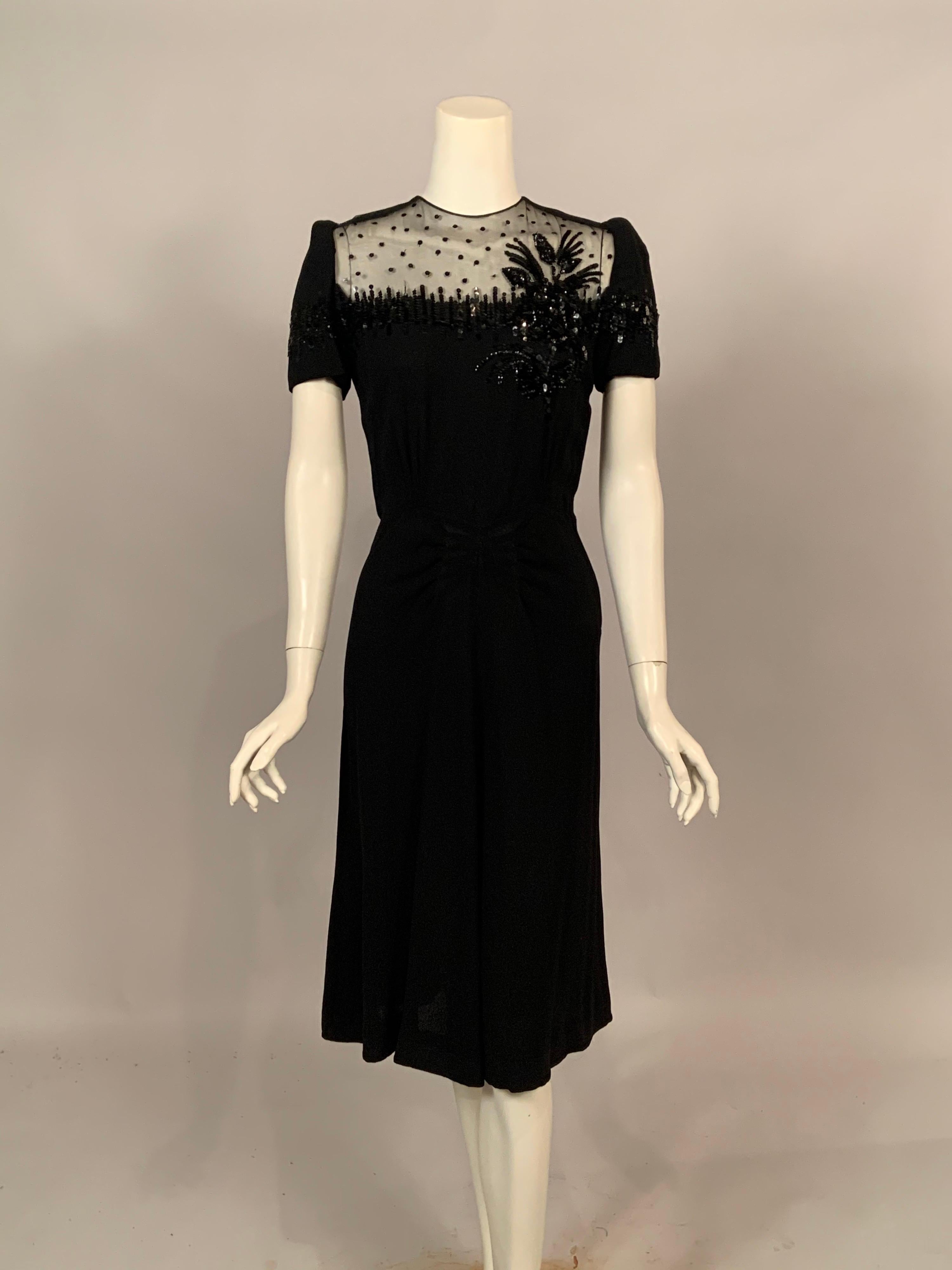 A truly glamorous black crepe dress with a sheer panel embellished with scattered sequins, vertical rows of  black sequins on the sleeves and bustline and a beautiful three dimensional floral corsage on the left side. The dress has three vertical