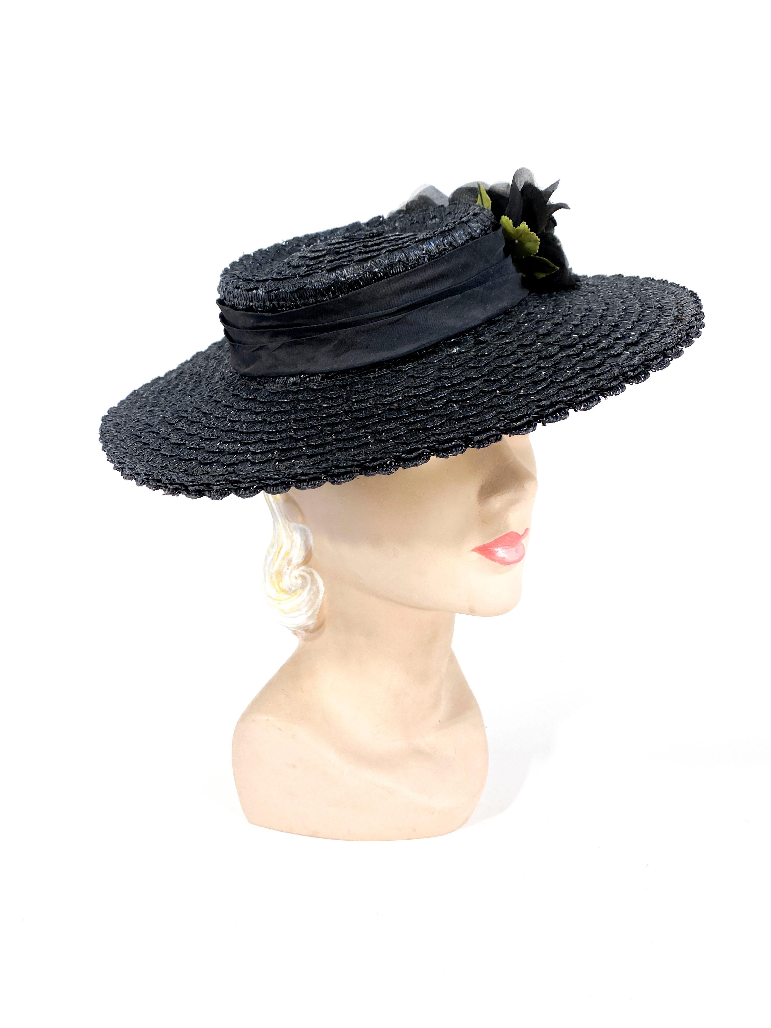 1940s black coated straw woven into many rows of spiraling scallops. The shallow crown is adorned with a gathered twill brand and a silk flower with green foliage. The black security band is worn on the back of the head to secure the hat.