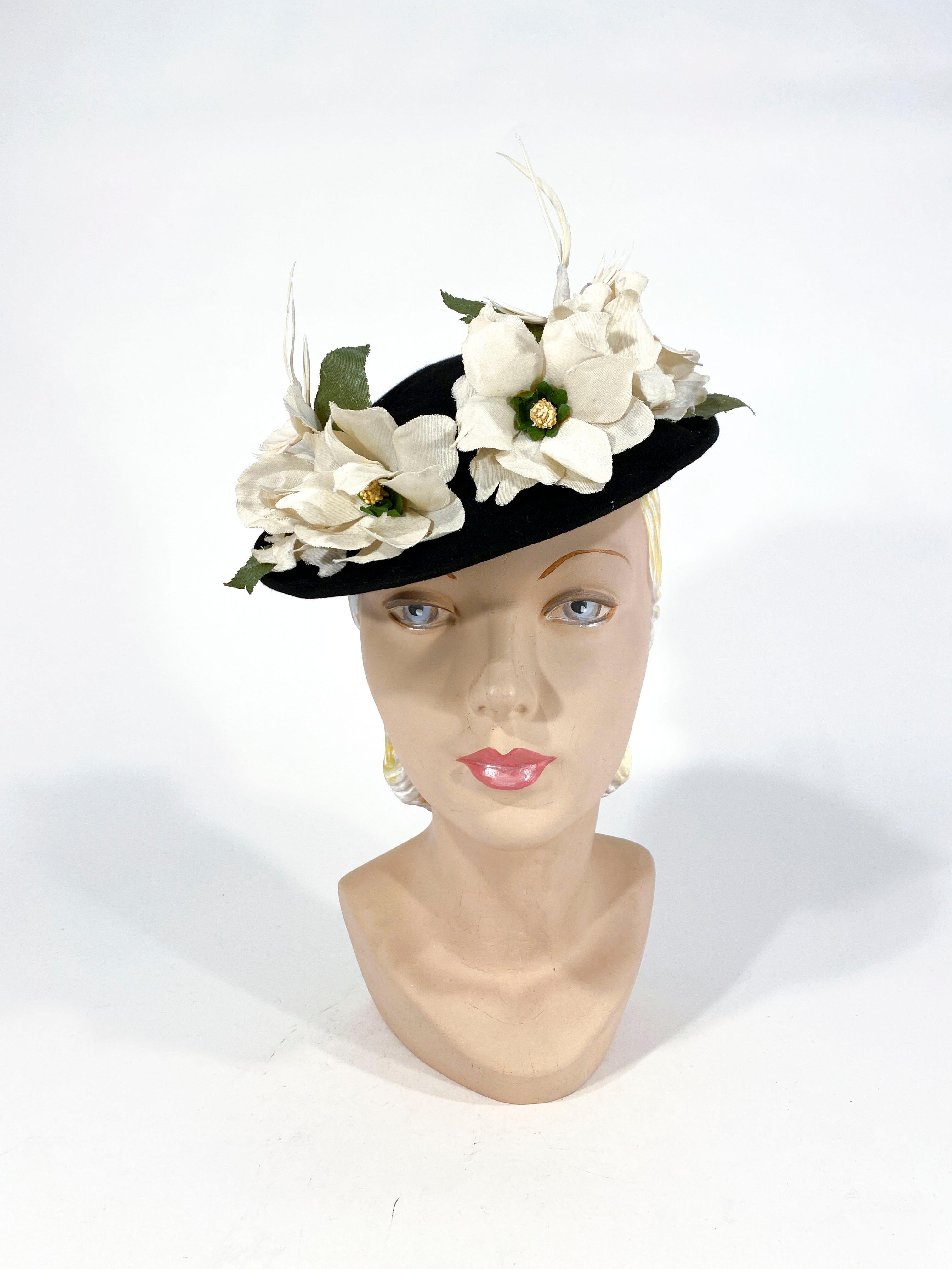 1940s black beaver fur felt toy hat decorated with handmade linen flowers, oil cloth leaves, and white feathers birds on each side. The interior has a security strap attached to secure the hat to the head. 