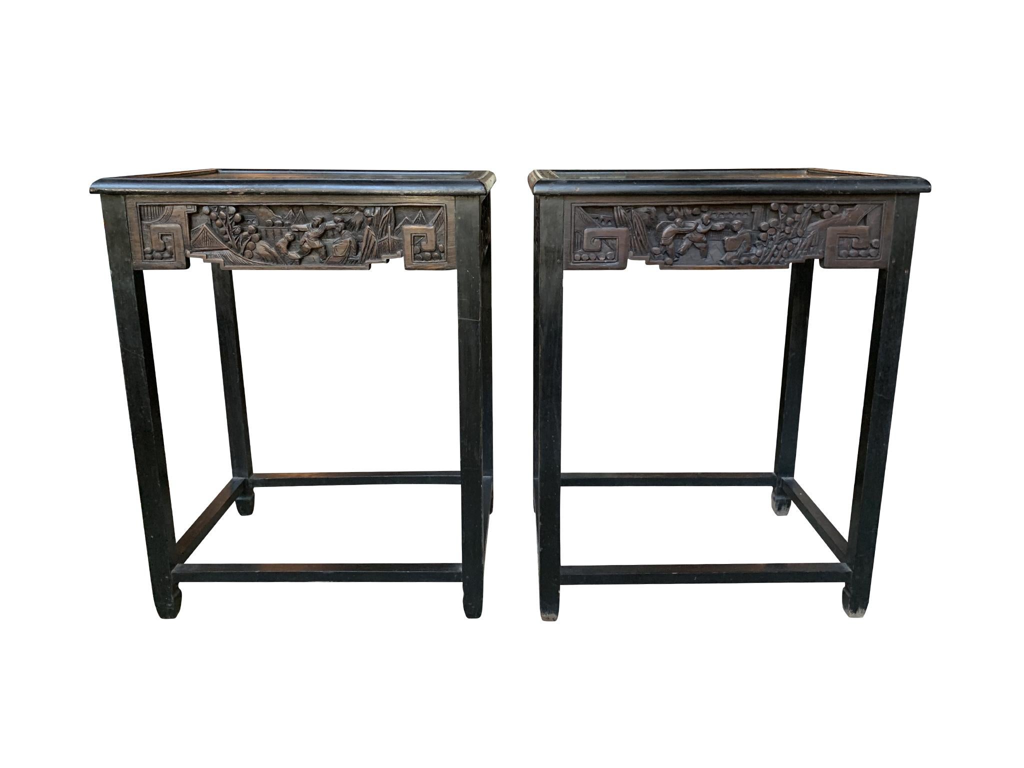 A pair of 1940s Chinese side tables, crafted with ebonized wood. The tables are remarkable for the hand carved, ornate frieze that runs along the skirt. A border of hand carved flowers frames the tabletop. The wood has a warm dark-brown tone. Its