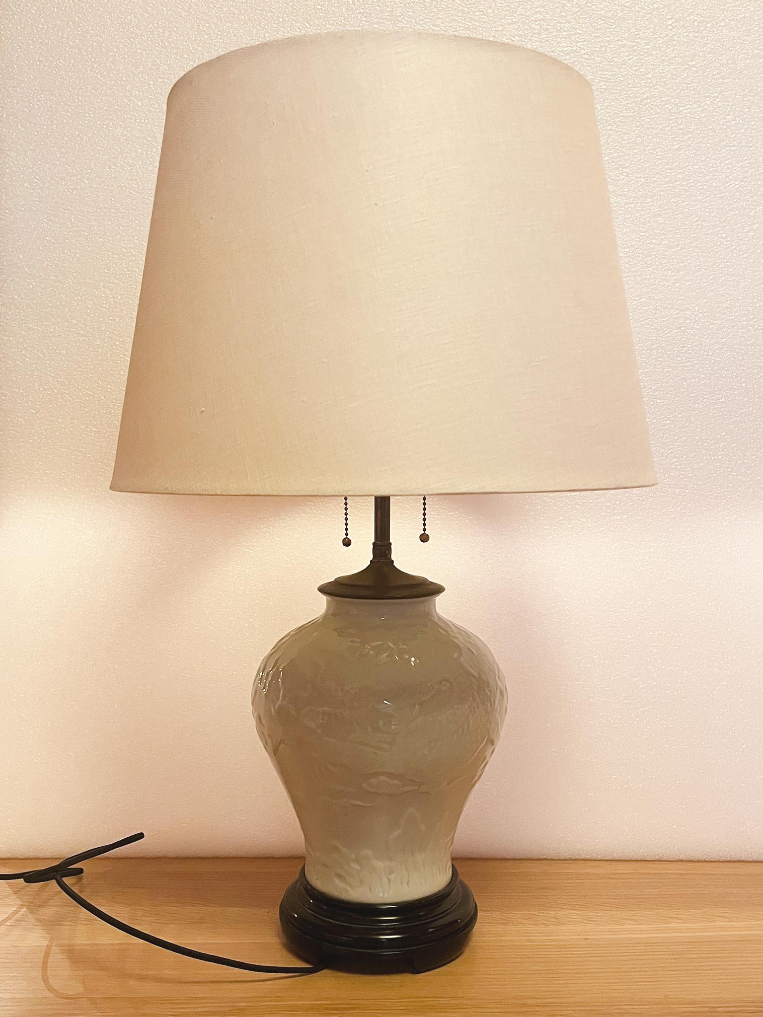 Fabriquée dans les années 1940, cette lampe de table chinoise se compose d'un corps d'urne en céramique à la glaçure blanche pâle sur un socle en bois noir. Un motif de dragon enroulé orne subtilement la surface. La lampe est dotée d'un groupe de
