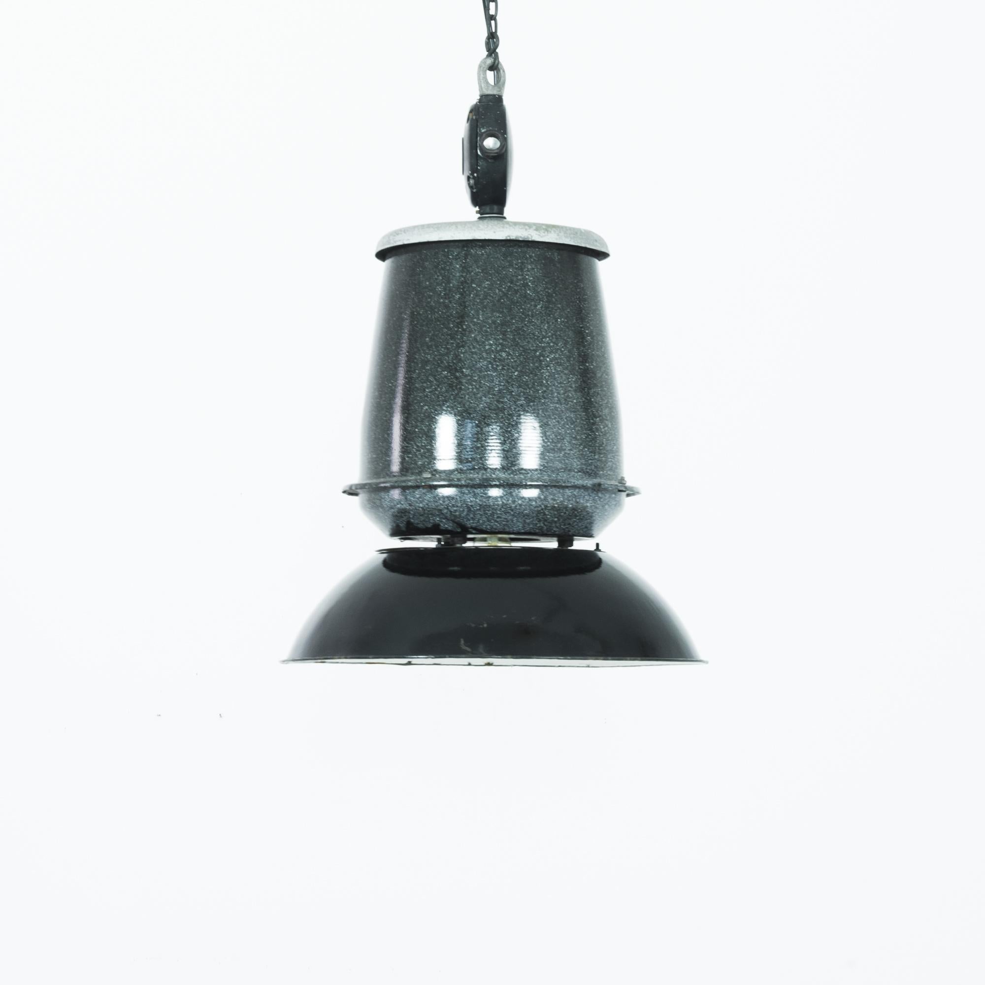 A pendant lamp from 1940s Czechia with an industrial silhouette. A broad metal lamp head hangs from a tapered cylinder; above, a round light fixture bears an original company logo. The speckled enamel of the pendent has a gentle gradient from black