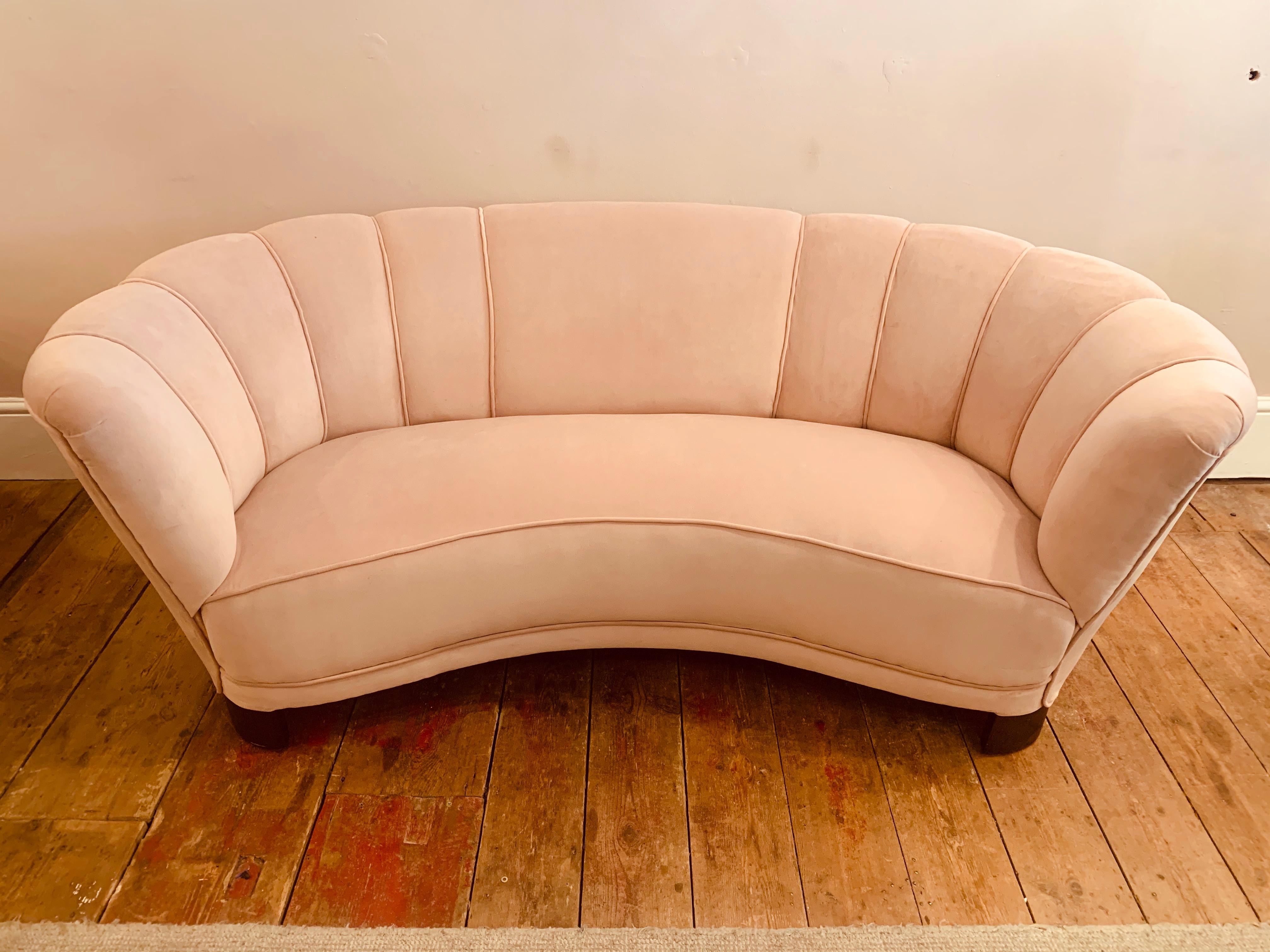 An elegant and imposing 1940s Danish banana-shaped scalloped sofa in the style of Viggo Boesen. The sofa has been newly reupholstered in a blush pink velvet fabric with edged feature piping at the seams and large brown curved wooden block