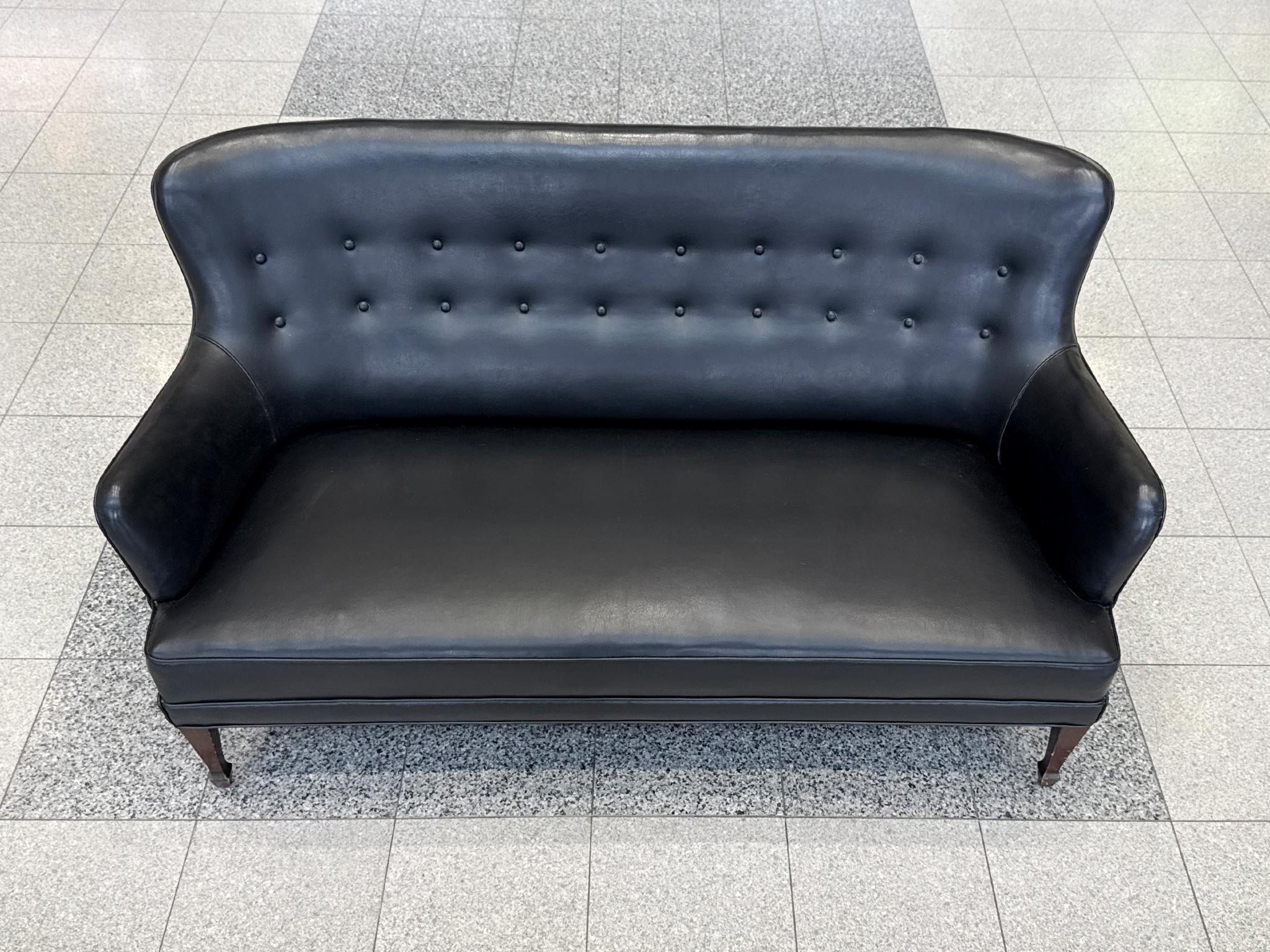Made in the 1940s, this black leather settee was designed and produced by the Danish cabinetmaker Georg Kofoed. Minimalist in design, the settee offers some subtle drama with its sloping arms, winged corners, and elegant button-tufted backrest. The