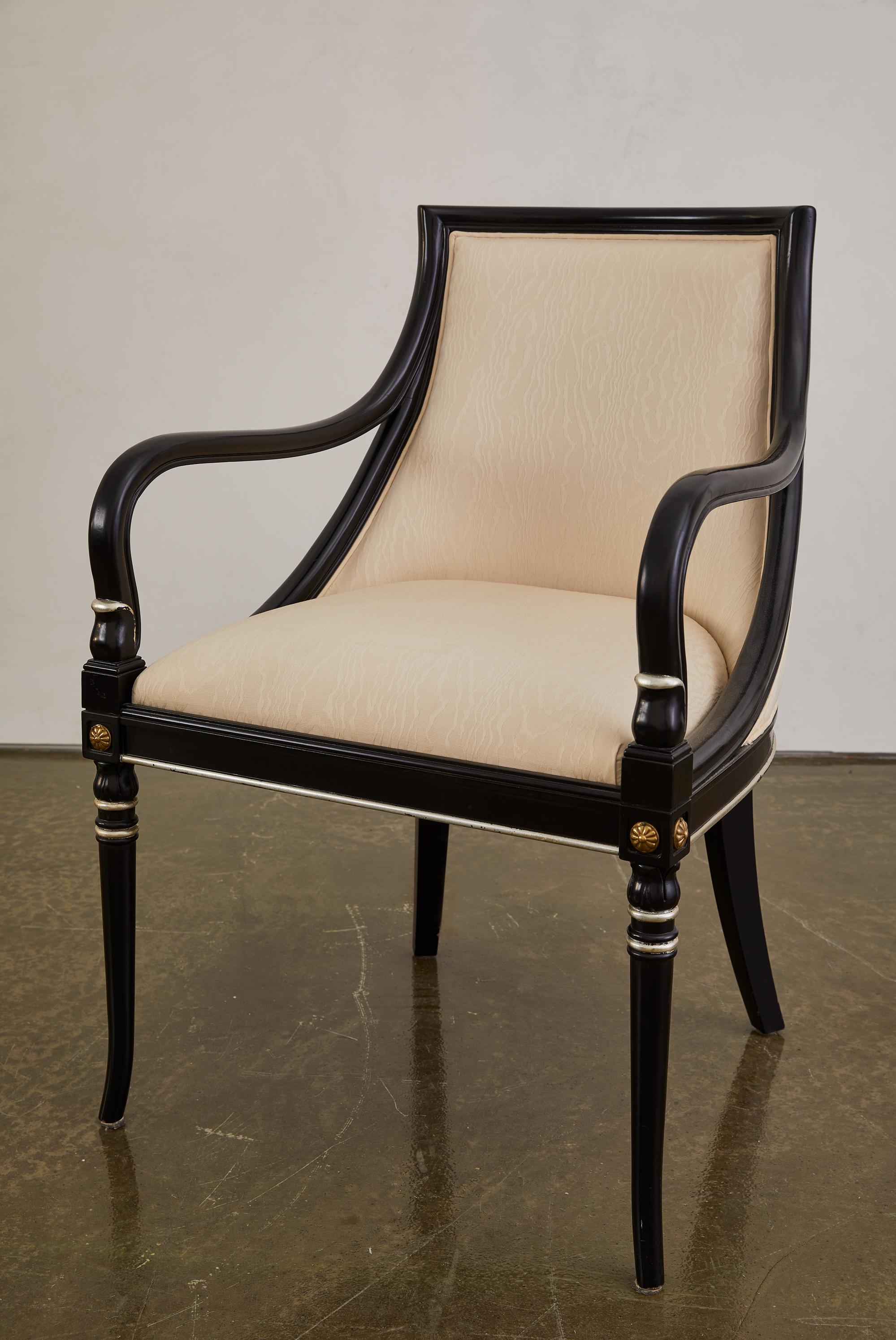 This striking pair of 1940s Regency Revival armchairs reflects the resurgence of neoclassical elegance in American design. Crafted with sculptural curved arms and an ebonized wood frame, these chairs are accented with gilt details and rosette