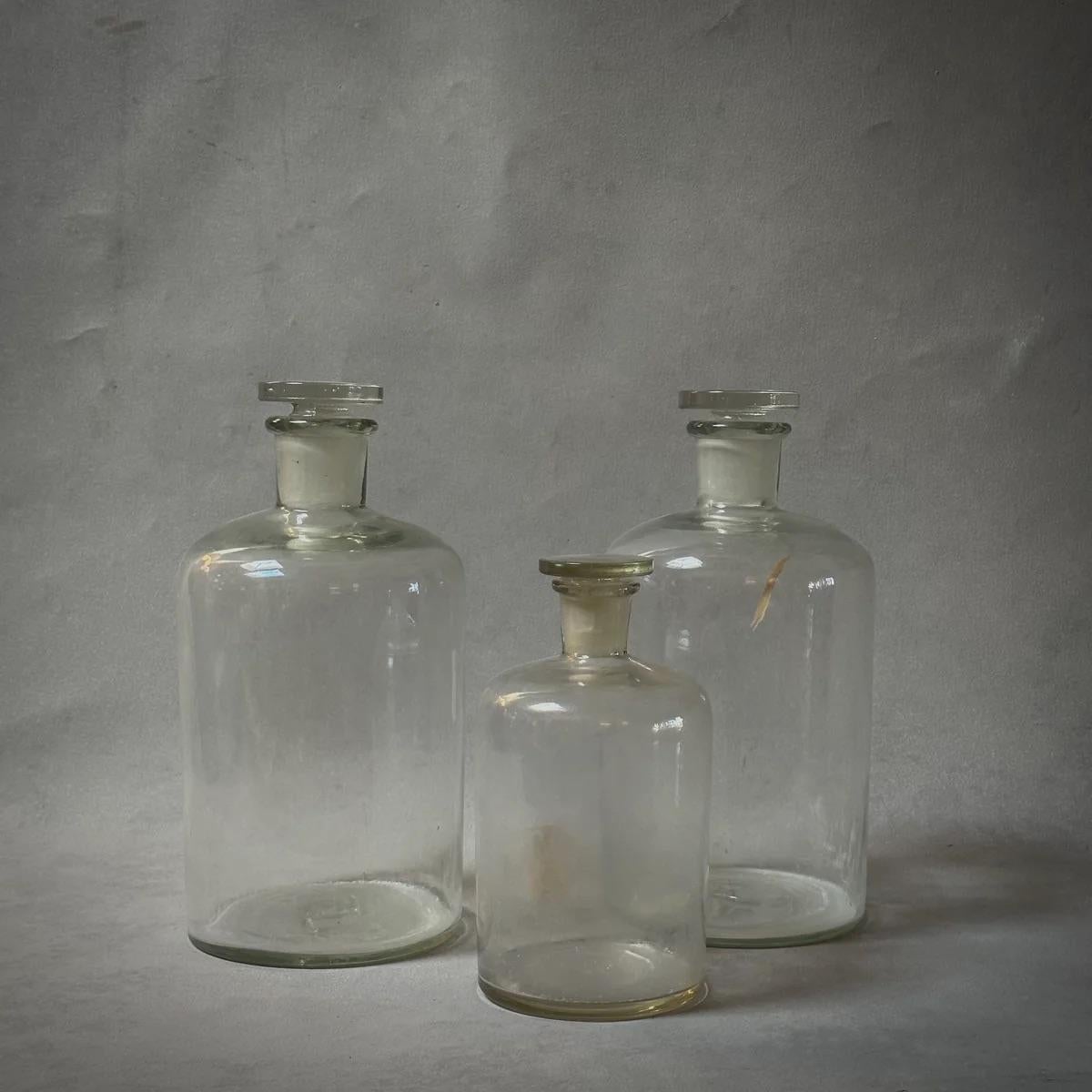 Diese Klarglasflaschen aus England aus der Mitte des Jahrhunderts, ca. 1940, werden mit runden, passenden Glasstopfen geliefert, die ihre schlichte, aber robuste Form perfekt ergänzen. Die Gläser haben ein angenehmes Gewicht und eine rustikale