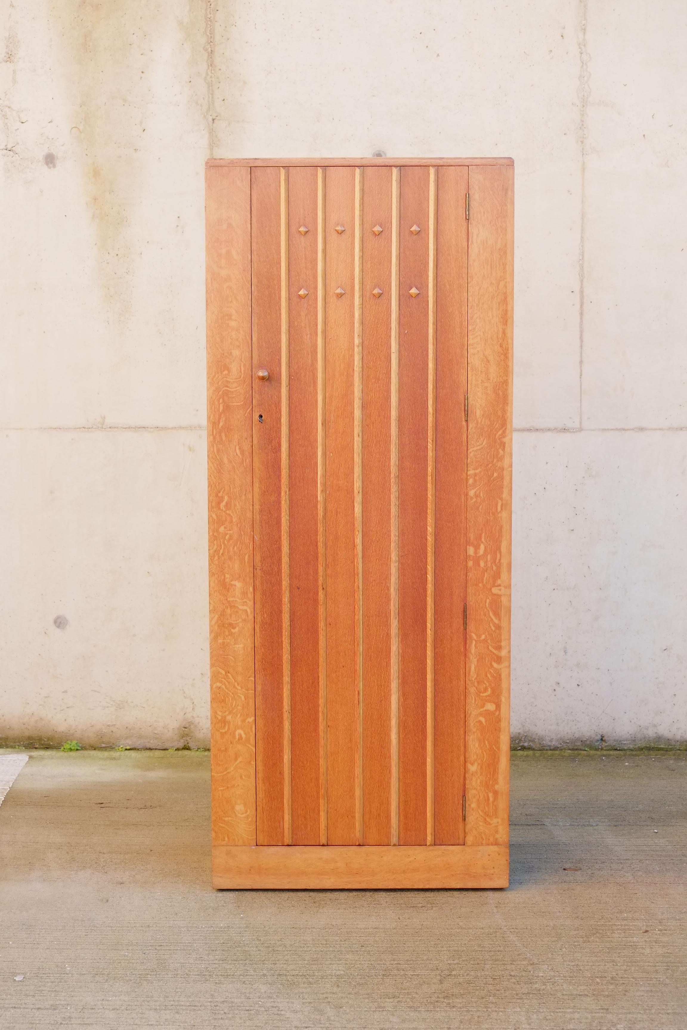 Une élégante armoire haute et étroite, fabriquée en Angleterre vers 1940. L'armoire est composée d'un lambris en chêne clair avec un magnifique panneau et des détails en forme de diamant sur la façade de la porte. A l'intérieur, il y a une seule