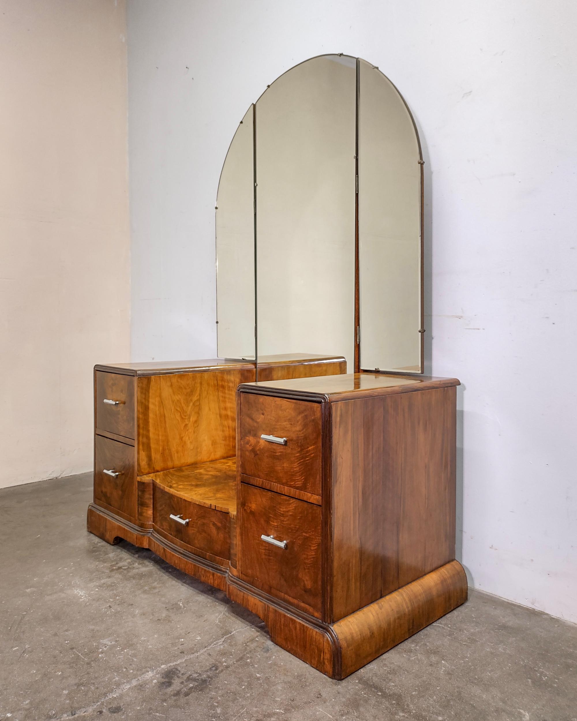 Beautiful art deco vanity with book-matched flame walnut veneer over solid wood. Triptych mirror with arched top, each side pivots outwards. Two deep drawers on either side and one in the center with original chrome hardware. Beautiful curvature on