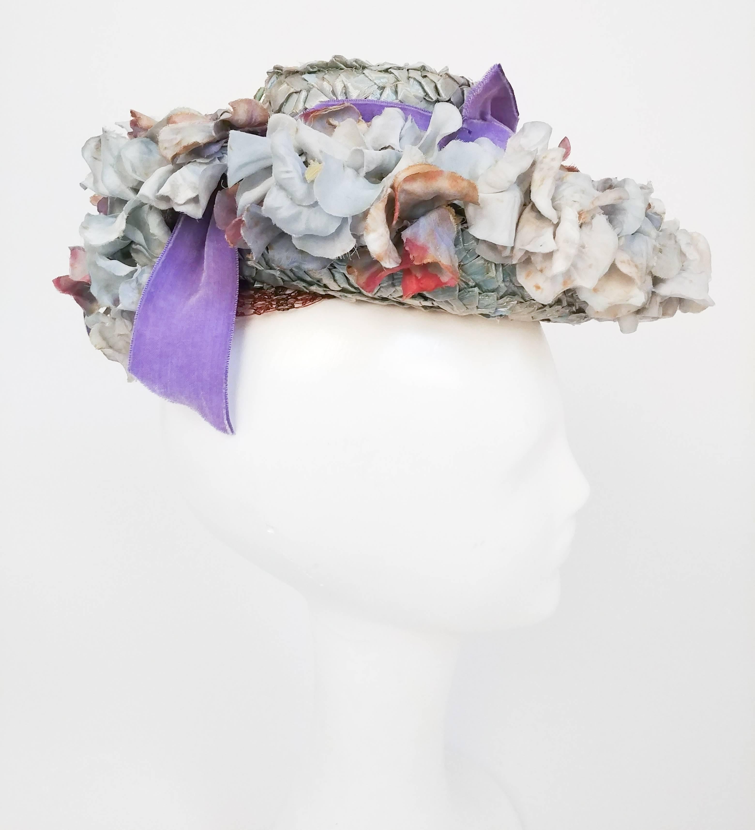 1940s Flower Hat w/ Lavender Velvet Ribbon. Raffia base hat with velvet ribbon, mesh net detail at crown, and decorative flowers all around. Held to head with elastic strap at back. 