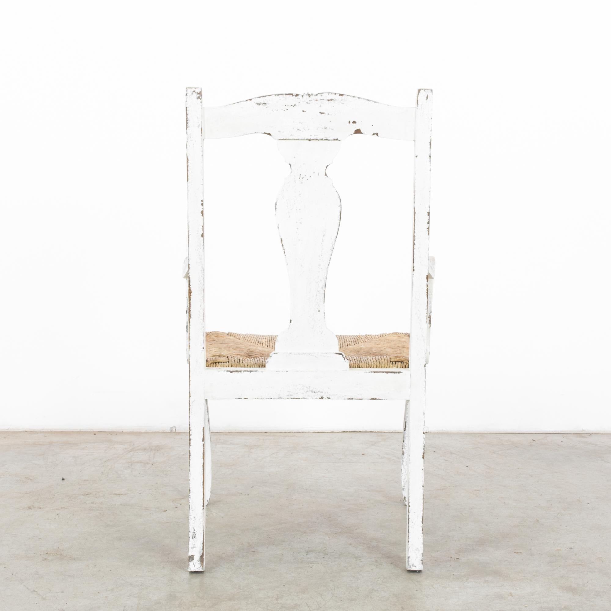 This wooden chair with a woven seat was made in France, circa 1940. The carved center splat and top rail enhance its rustic charm, while the curves of the armrests and cabriole legs offer a graceful elegance. Beautifully weathered with a white