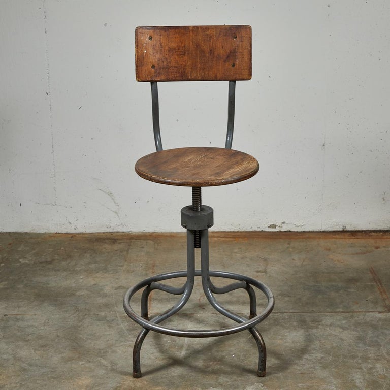 1940s French industrial wooden swivel stool with adjustable steel base. Featuring a flat circular seat and curved rectangular back, the chair sits on a spider base with four enjoined legs inscribed by a circular foot-rest. The modernist, utilitarian
