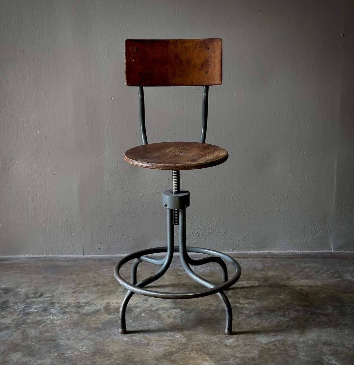 This industrial swivel stool perfectly embodies the era's appreciation for functionality fused with style. Its wooden seat and curved rectangular backrest are mounted on an adjustable steel spider base, complete with a circular footrest. The