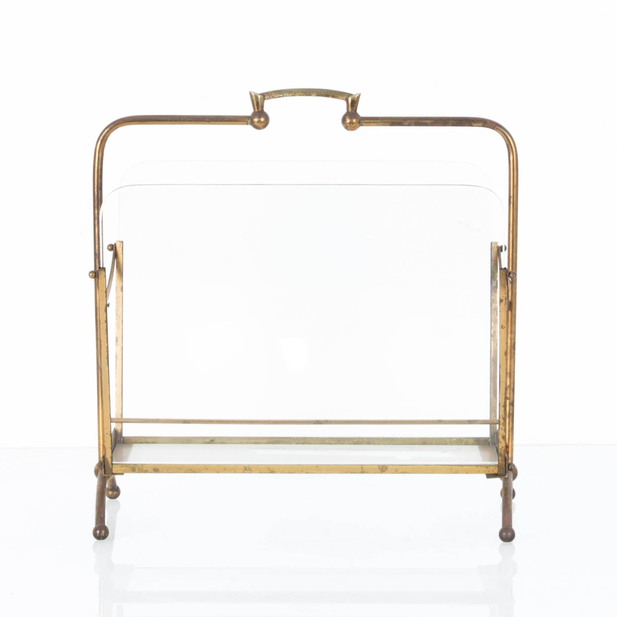 This glass and metal newspaper stand was made in France, circa 1940. The rounded edges and arcs of the legs and sides give this rack a pleasing form, while details such as the handle and ball feet add charm. Finding your newspapers and magazines