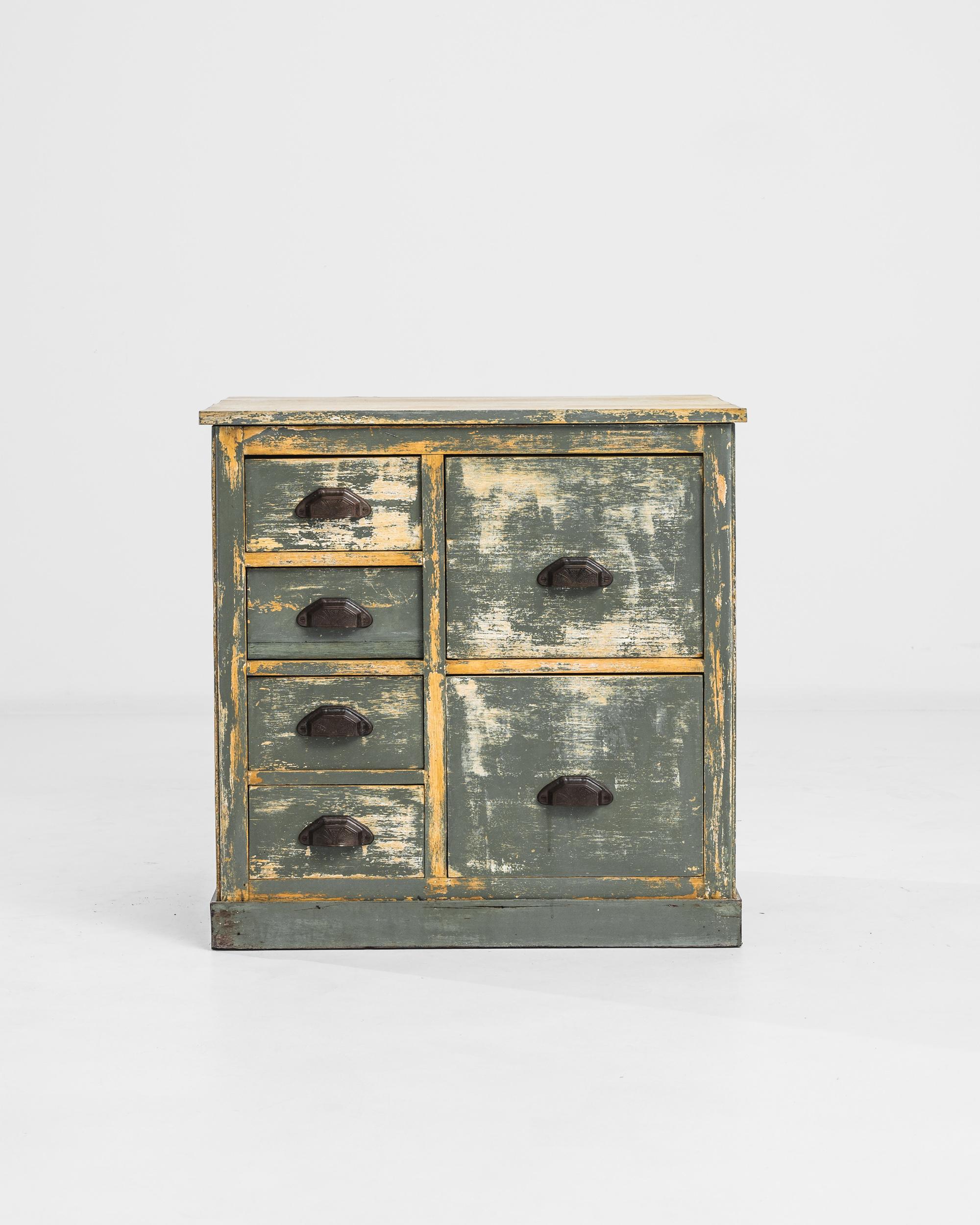 A dignified wooden chest of drawers with an attractive distressed finish. This vintage piece was built in 1940s France: deep, filing cabinet style drawers speak to a practical disposition, while ornate metal cup pulls, embossed with a pattern