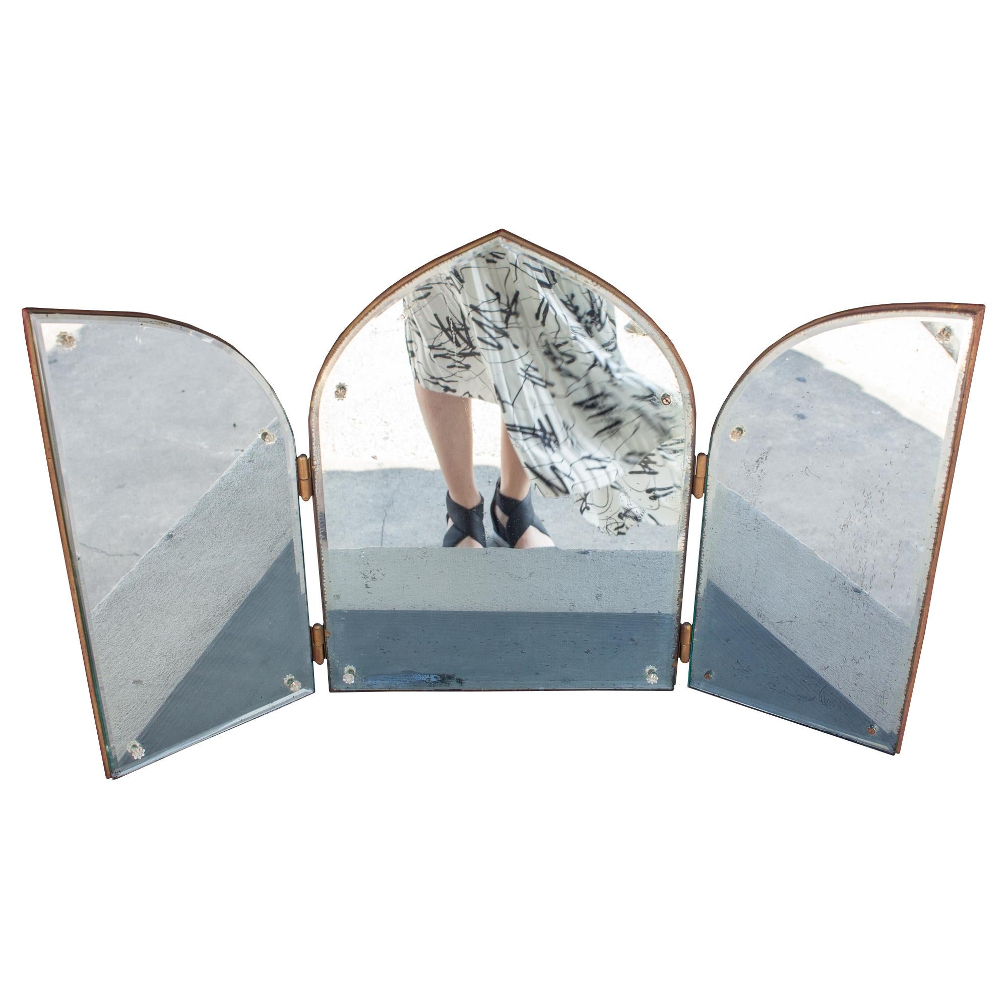 This 1940s triptych mirror has three panels, hinged together. The shape is arched and features a beveled edge on all three mirror panels with clear decorative caps on the screws (9 of 12 are intact). The back of the mirror is wood and has been