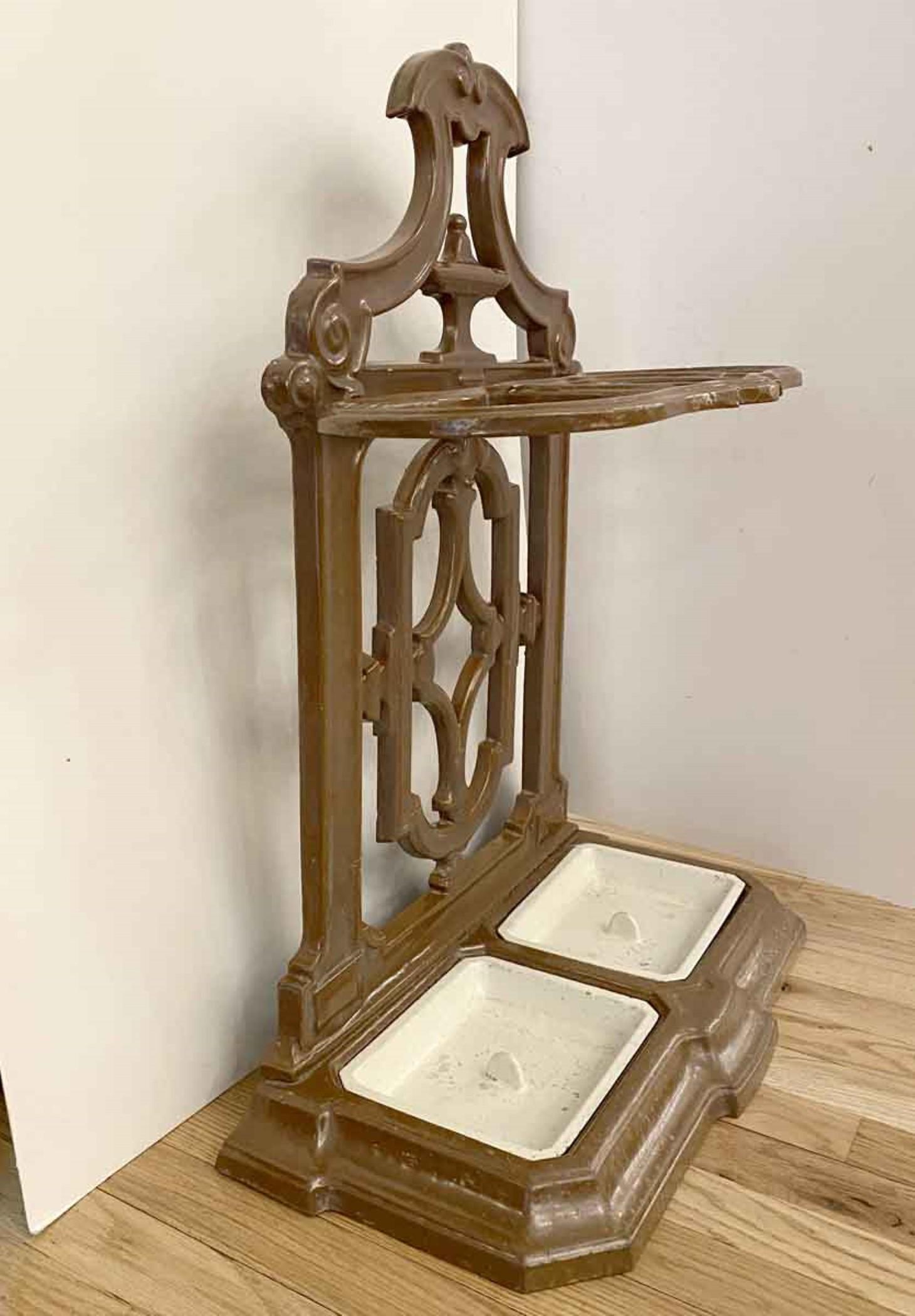 This 1940s ornate umbrella stand is well designed for wet umbrellas. It is a sturdy weight being made of enameled cast iron. The enamel is brown, and it has two white enamel trays to catch water drippings. There are three ring-like designs that will