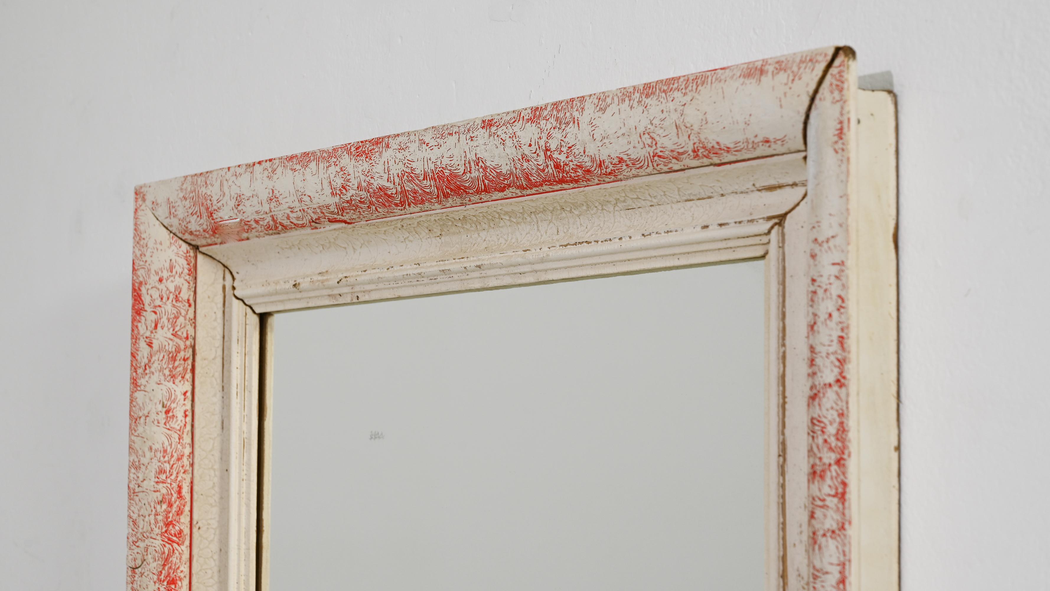 This 1940s French wood mirror exudes a serene elegance with its white patinated finish that gracefully frames the reflective glass. The distressed red undertones provide a subtle hint of color, suggesting a storied past and a rich history. This