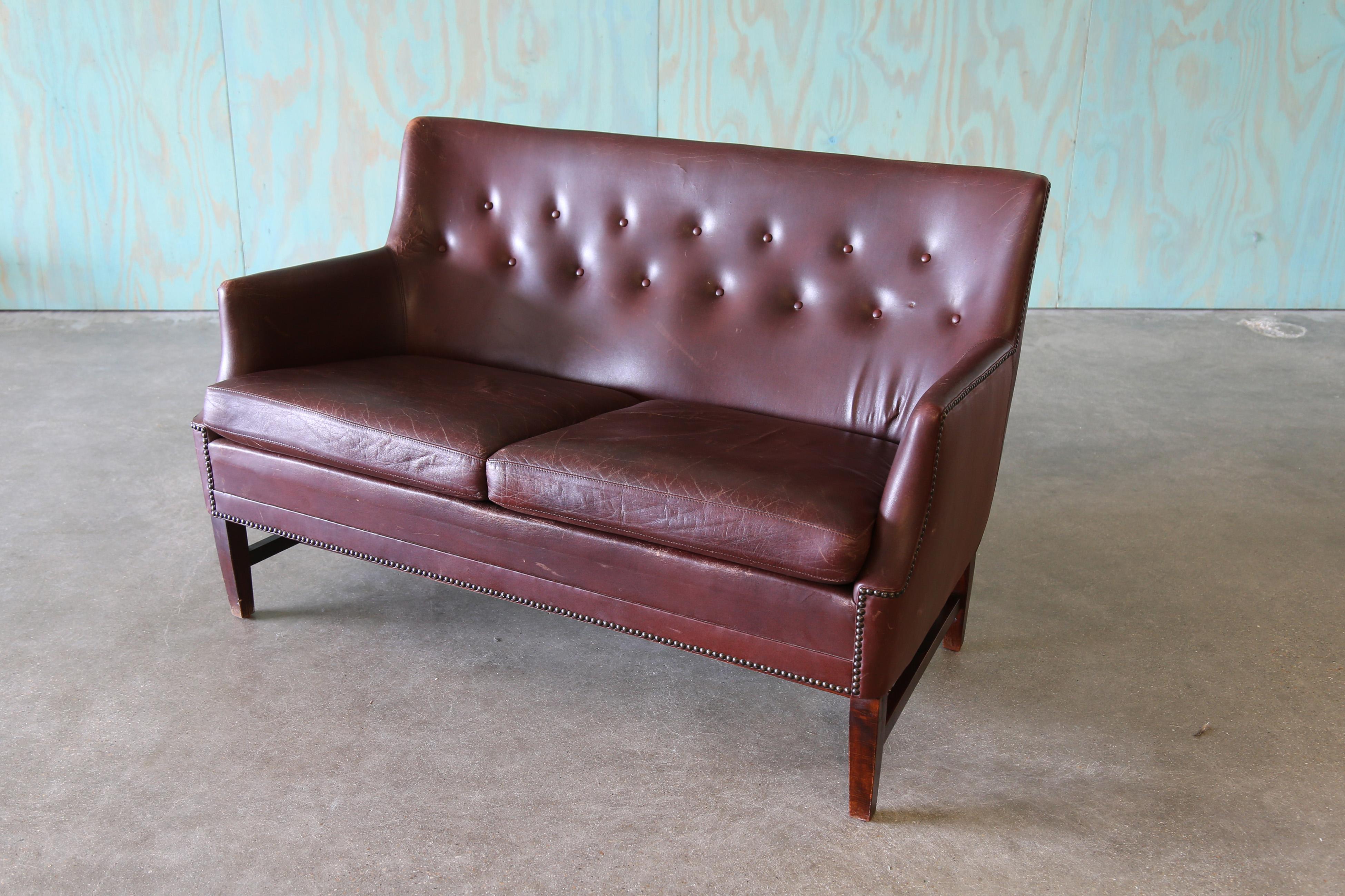 A diminutive danish brown leather sofa / couch in the manner of Fritz Henningsen. The button tufting accentuates the curved back. Similar in styling to Finn Juhl, and Flemming Lassen this settee could work in a formal library to a minimalist danish