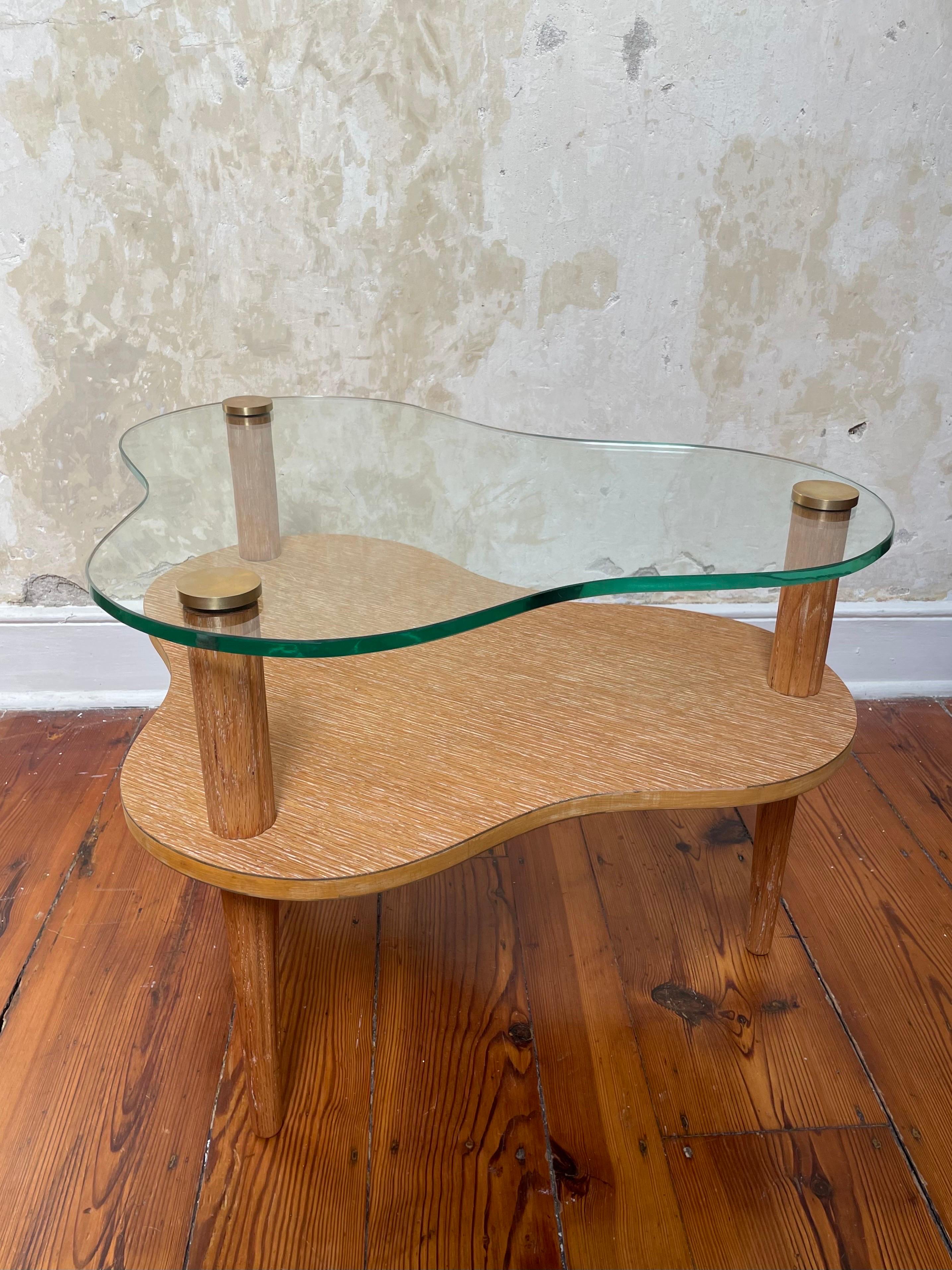 A glass and cerused oak 2 tier cloud shaped accent table with carved tripod legs and brass caps. Designed by Gilbert Rhode for Herman Miller for the early 1940s Paldao Group. The forms and materials of this line paid homage to the work of the