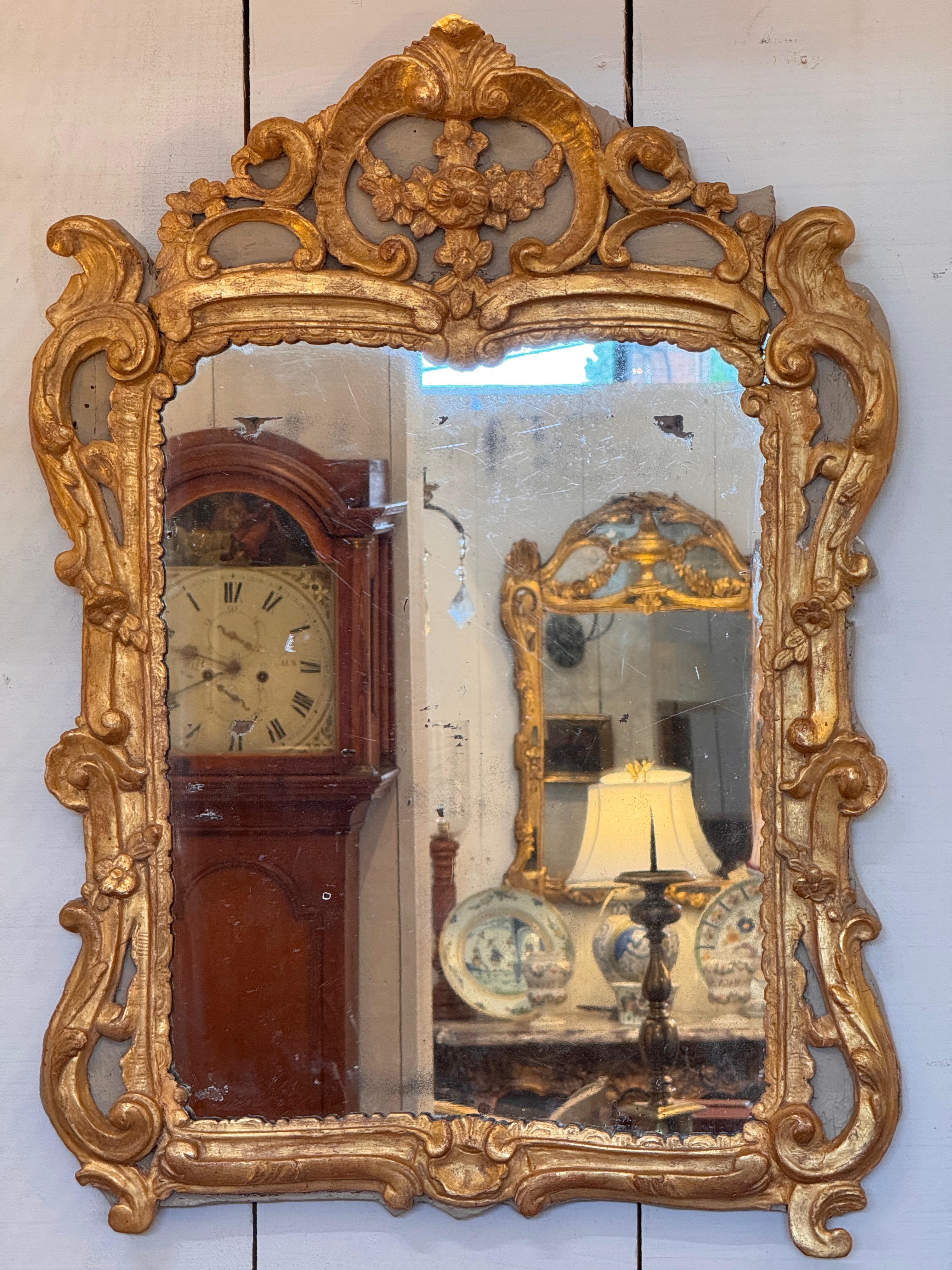 Vintage giltwood mirror with an ornate pierced crest featuring floral and scroll motifs. The frame displays deep carving and shaped edges with classic rococo influence, highlighted by soft wear to the gilding that enhances its age and character.
