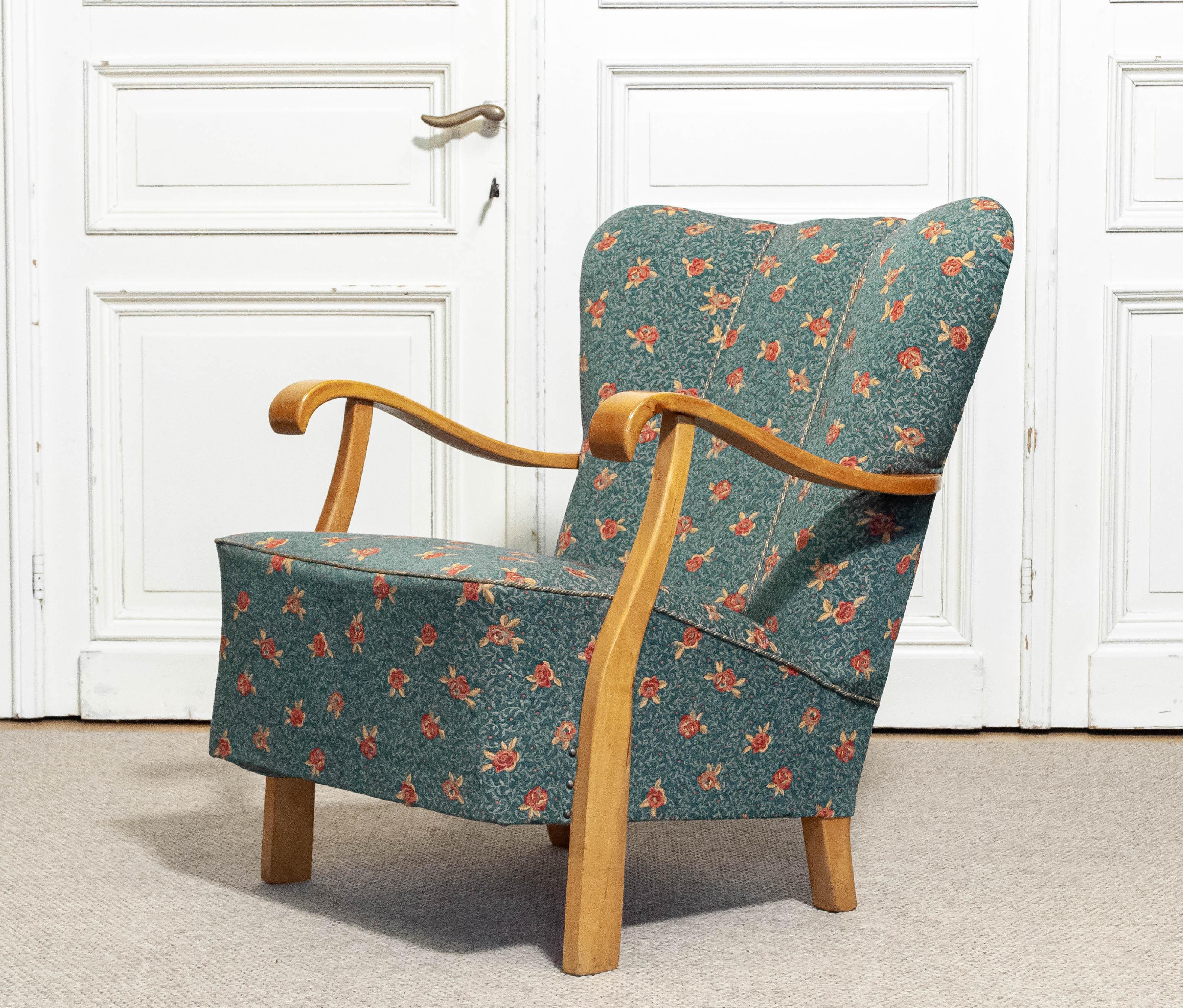 Si tratta di una poltrona da salotto rivestita in tessuto jacquard verde con un motivo floreale, risalente alla metà del XX secolo. La sedia presenta una solida struttura in legno con braccioli elegantemente curvati, che evidenziano l'artigianato