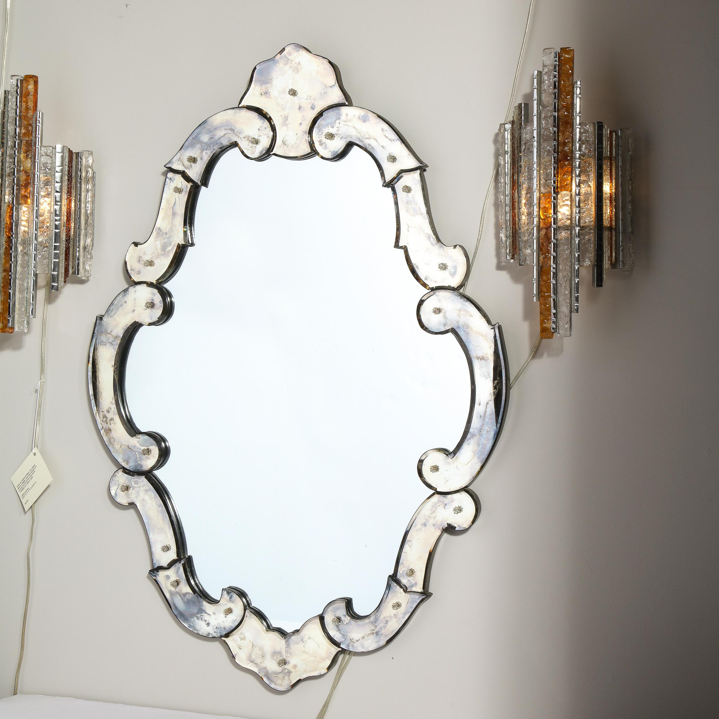 This elegant early Mid-Century Modern/ Hollywood cartouche wall mirror was realized in the United States, circa 1945. It features a cartouche form composed of an abundance of stylized demilune smoked and antiqued mirrored pieces that create a