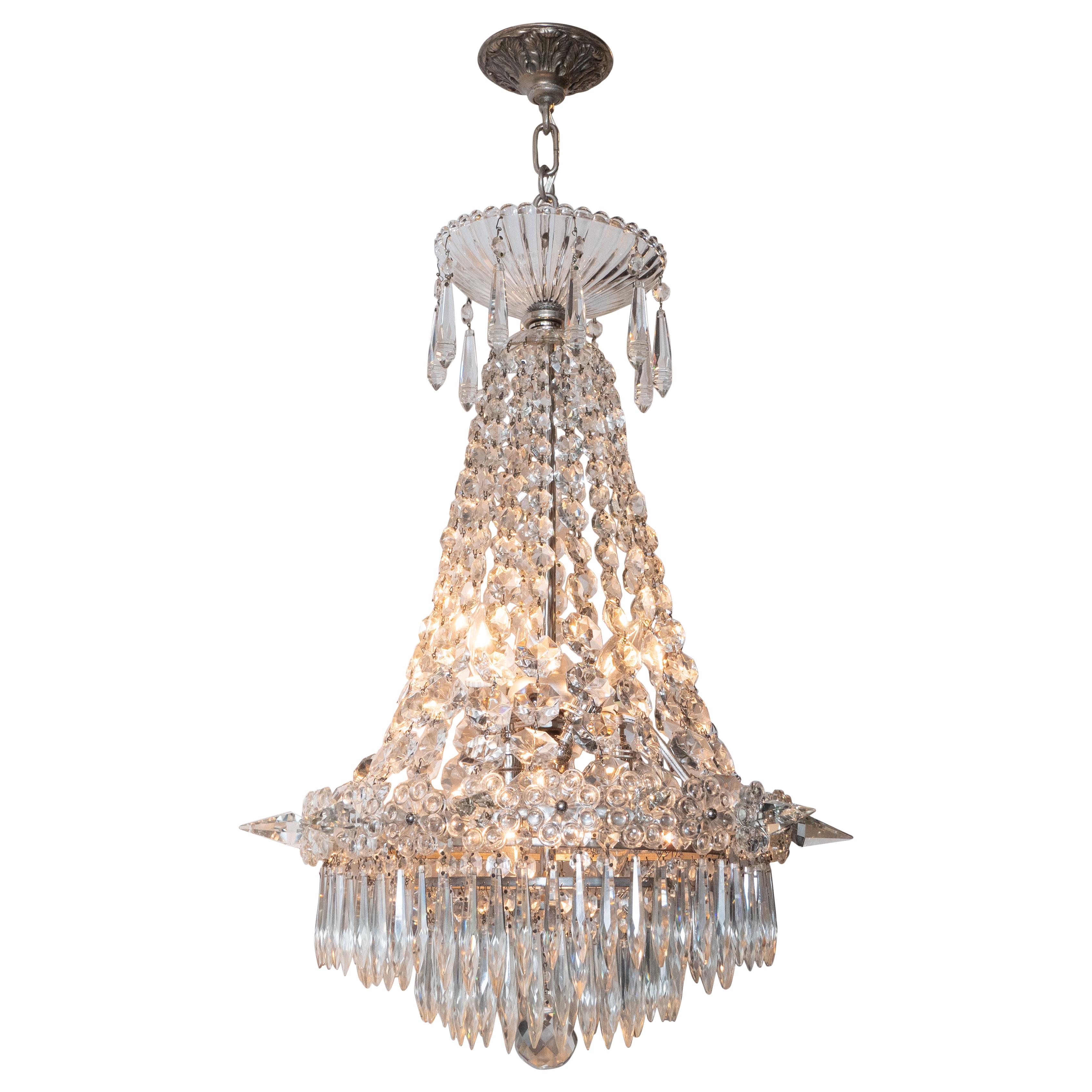 This elegant four tier Hollywood Regency chandelier was realized in the United States, circa 1940. It features an abundance of fine crystals suspended from a silvered bronze frame. From a circular silvered bronze canopy- decorated with stylized