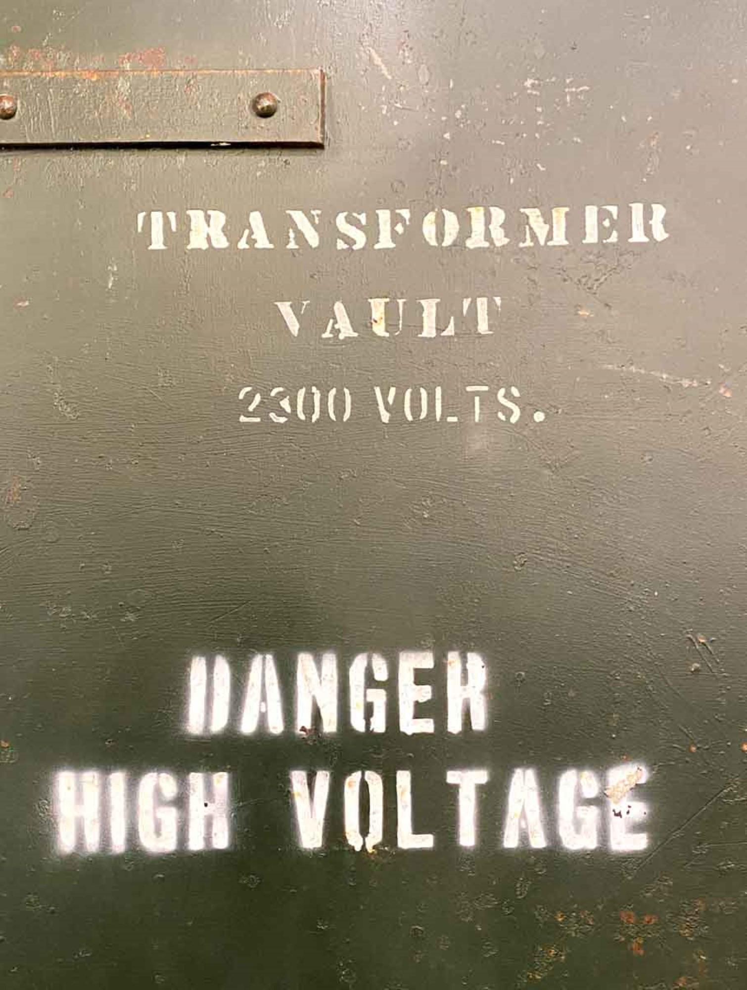 This green steel door reads transformer vault 2300 Volts. It is stenciled danger high voltage. The three strap hinges are each 6 inches tall, and the middle one currently does not move, probably from rust. The door is enforced with steel pieces