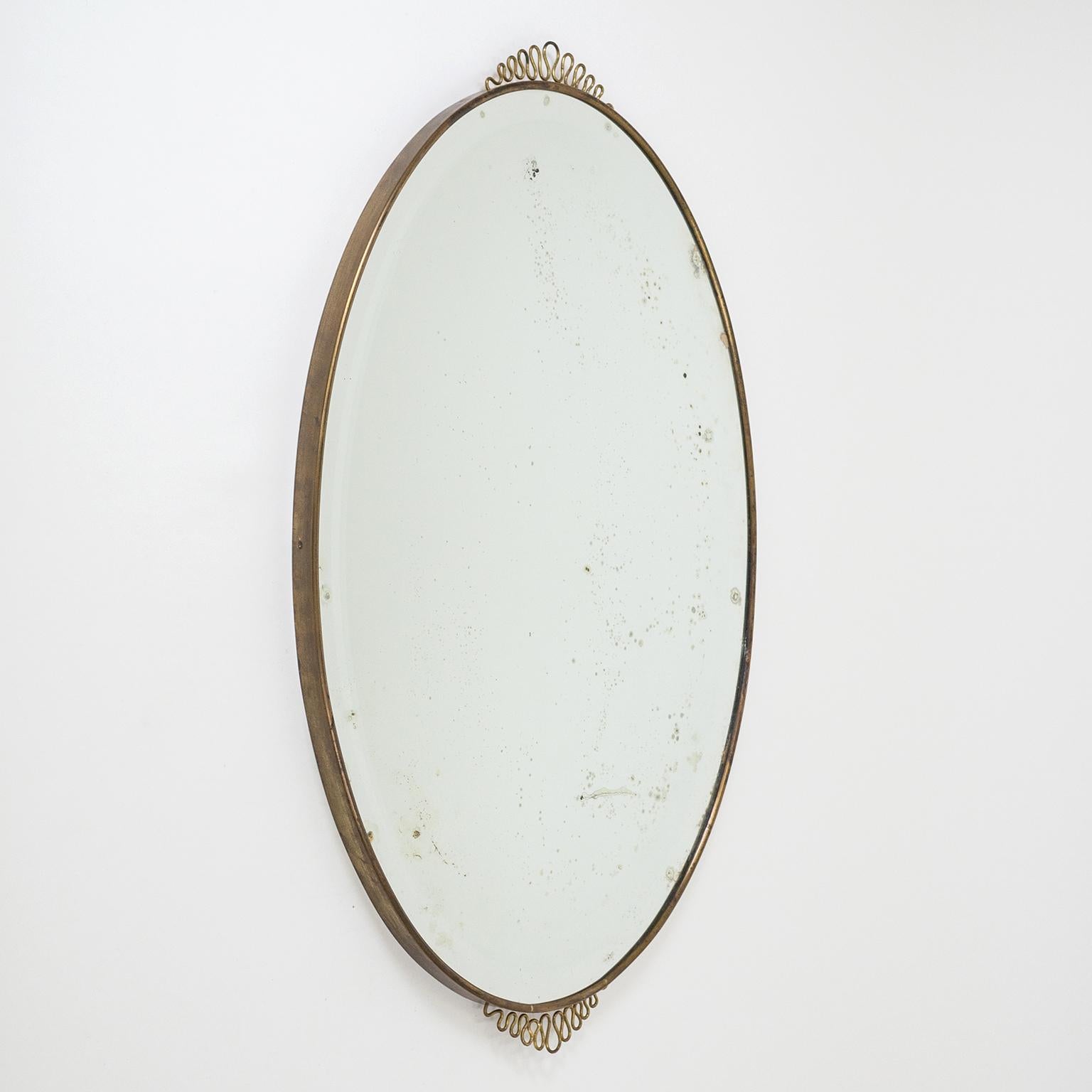Lovely oval Italian brass mirror from the 1940s in the style of Gio Ponti. Adorned with two undulating brass finials. There is a charming patina on the brass and heavy wear on the original faceted mirror with blind spots and corrosion to the silver