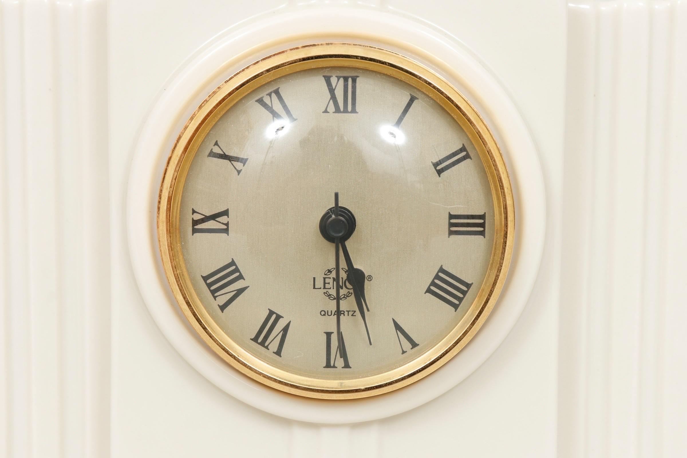 An Art Deco style porcelain mantel clock, made by Lenox for their Timely Traditions collection. White porcelain decorated with beveled columns support a round gilt/brass framed clock face with Roman numerals, marked “Lenox” with a wreath and