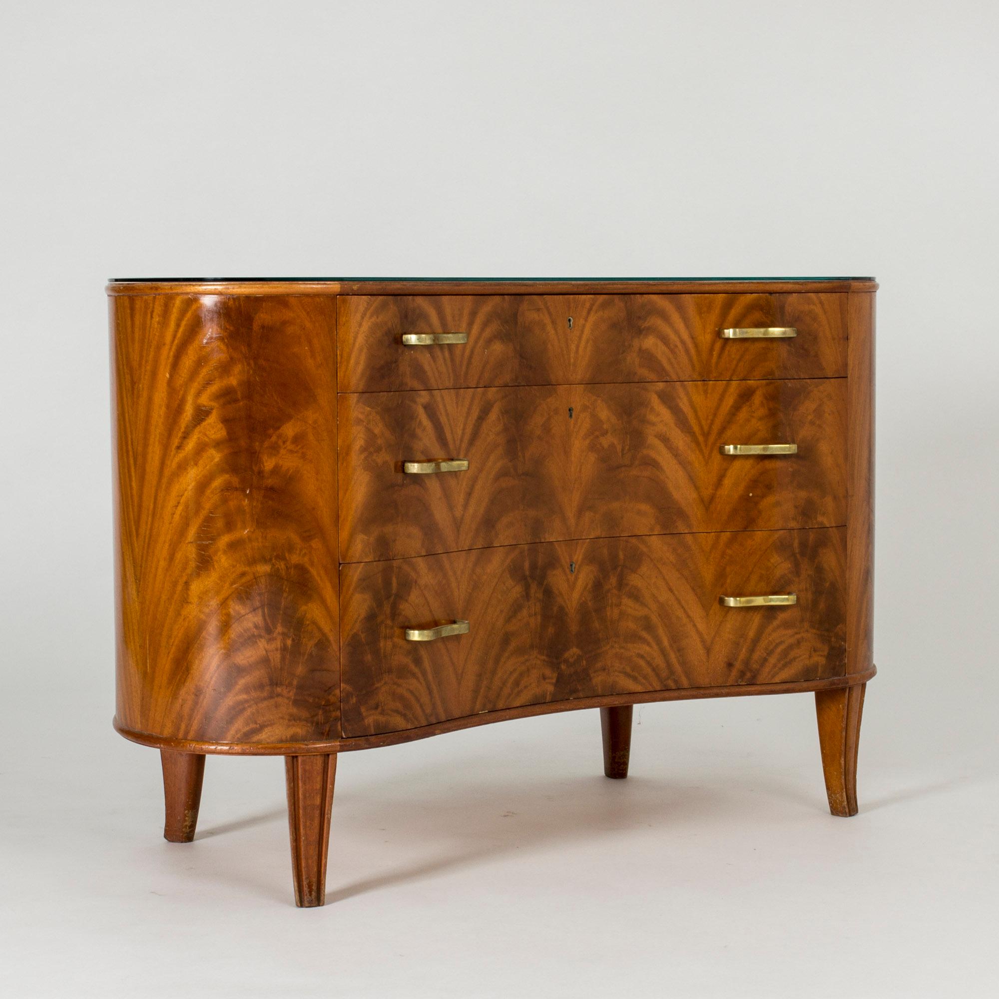 Beautiful, kidney-shaped mahogany chest of drawers from Bodafors, with elegant curved brass handles and green-tinted glass on the top. The veneer has been arranged to create a dramatic effect which contrasts nicely with the gentle design. Great