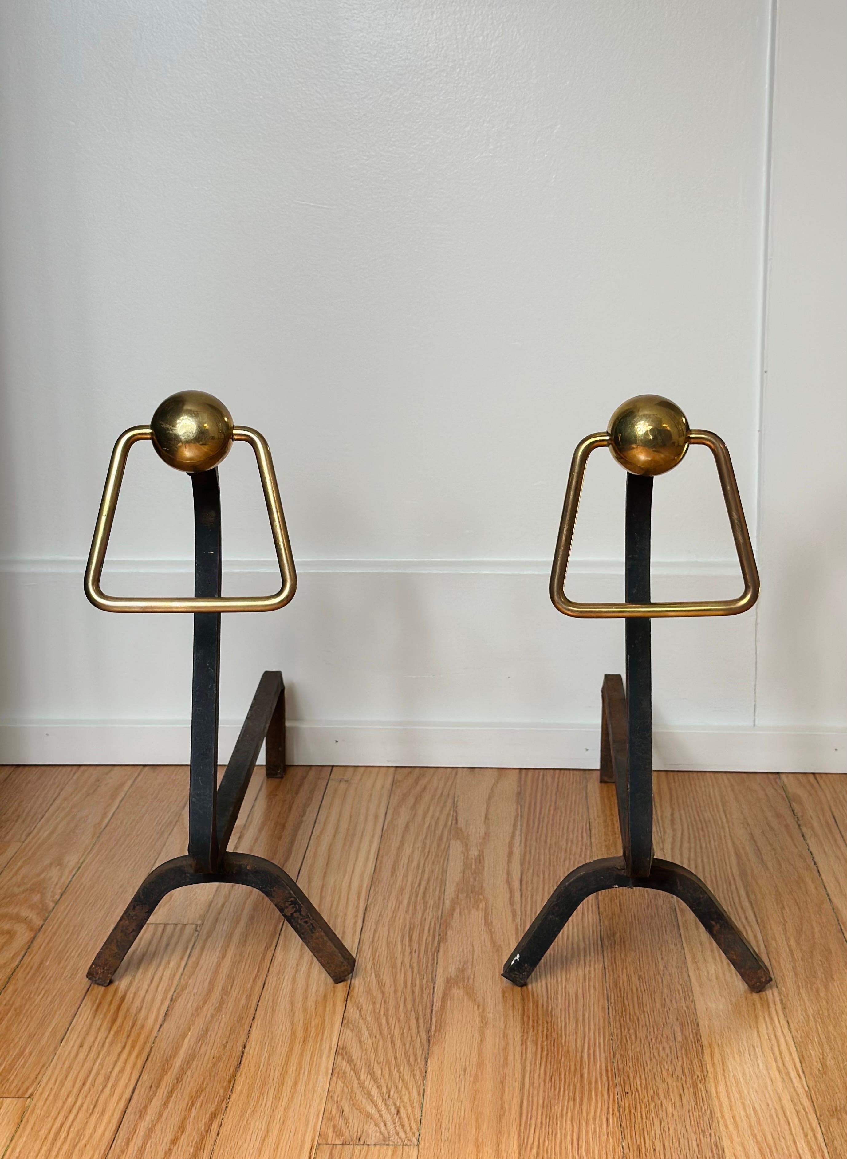 We are very pleased to offer a pair of Modernist andirons, circa the 1940s.  Crafted with meticulous attention to detail, these andirons seamlessly merge wrought iron and brass elements.  The wrought iron is shaped into a sleek and streamlined