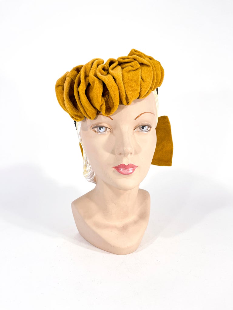 1940s mustard gold velour hand-molded cocktail hat with a graduated ruffle decorating the front edge of the hat and a back panel finished with an enlarged matching bow. The box is attached to an olive green security back band to secure the hat to