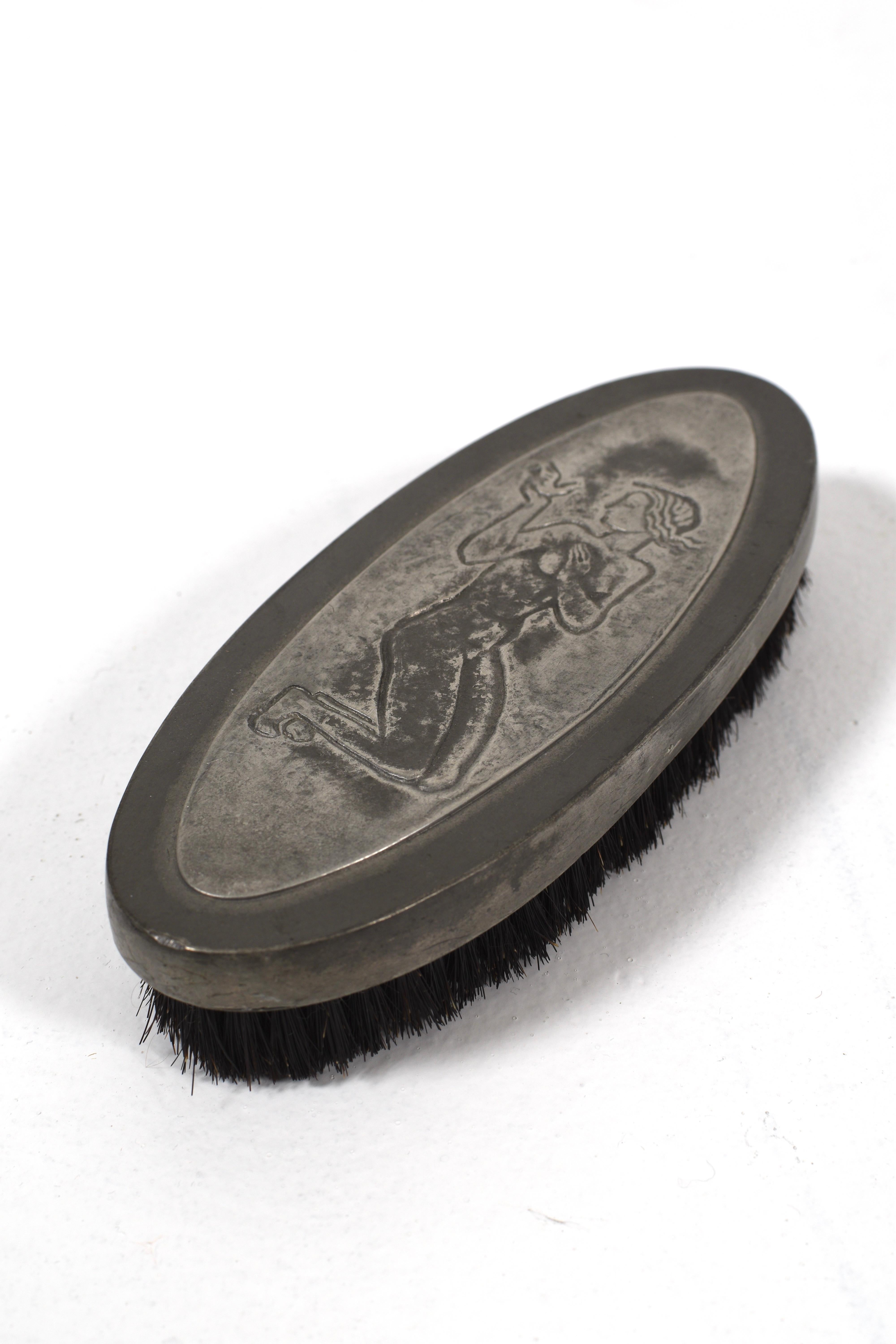 This pewter clothes brush from 1940 combines practical function with elegant design. The top is adorned with a detailed relief of a kneeling woman holding a bird, crafted in a refined Art Deco style typical of the period.

The oval-shaped body fits