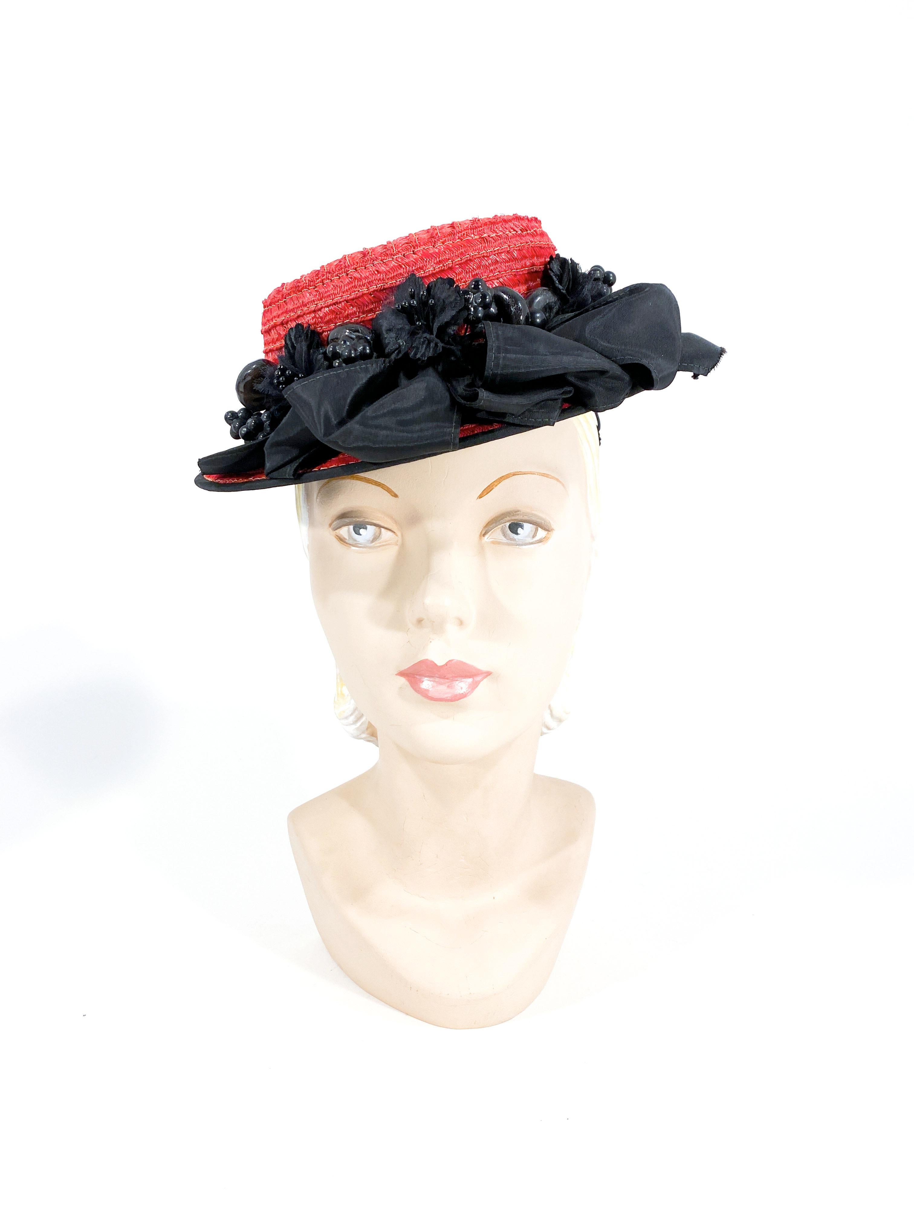 1940s red coated and woven straw toy pork pie hat. The short brim is decorated with a black taffeta trim, large looped taffeta ruffles, black lacquered wooden fruit, a narrow band, and silk hand-cut flowers. The interior hat band has an attached