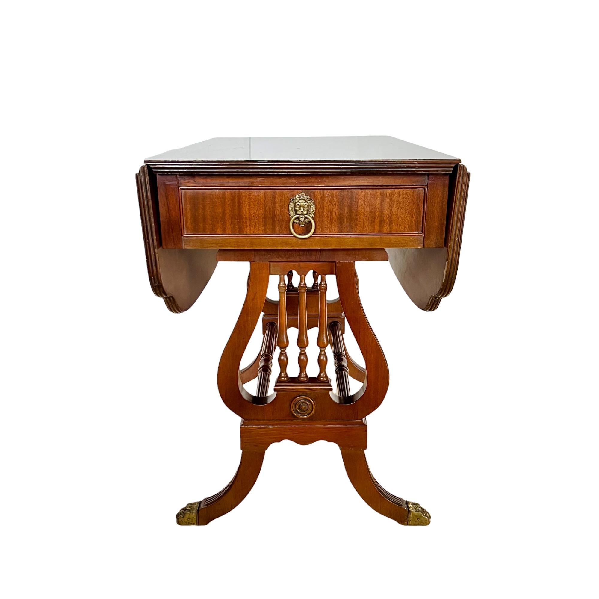 A vintage English Regency style mahogany drop-leaf accent or breakfast table with single drawer. Featuring a rectangular top surface with rounded cut-out corner detail, dual carved and turned stretchers between harp form lyre ends, channeled saber
