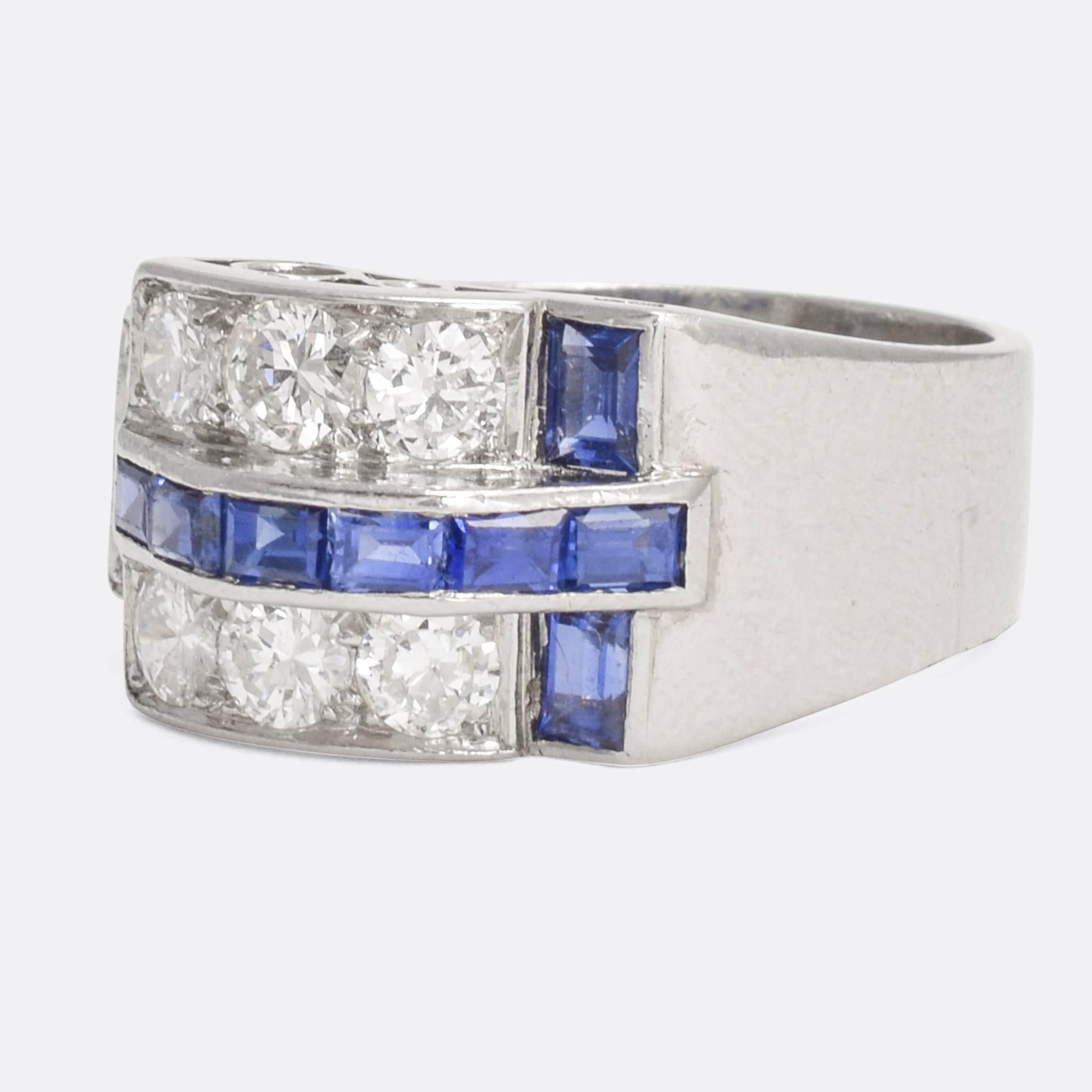 This cool Retro era ring was made in the 1940s. It's big and bold with a stong modernist vibe; a mid-20th Century take on the buckle ring, set with vibrant blue sapphires and brilliant cut diamonds. The gallery features fine scrolled openwork,