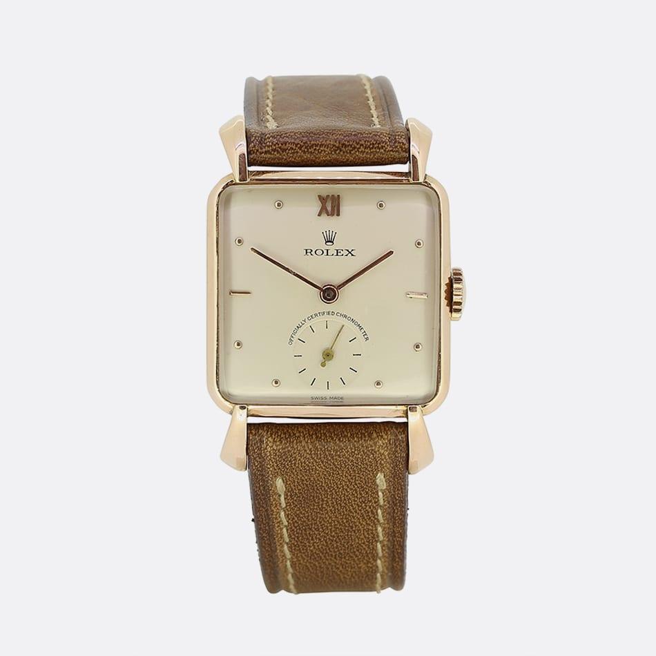 This is a 1940s 18ct rose gold unisex Rolex watch. The dial is a cream colour with gold hands and gold hour indices. The strap is brown leather strap with a gold buckle. The watch features an 18ct rose gold case with fancy lugs. Additionally, the
