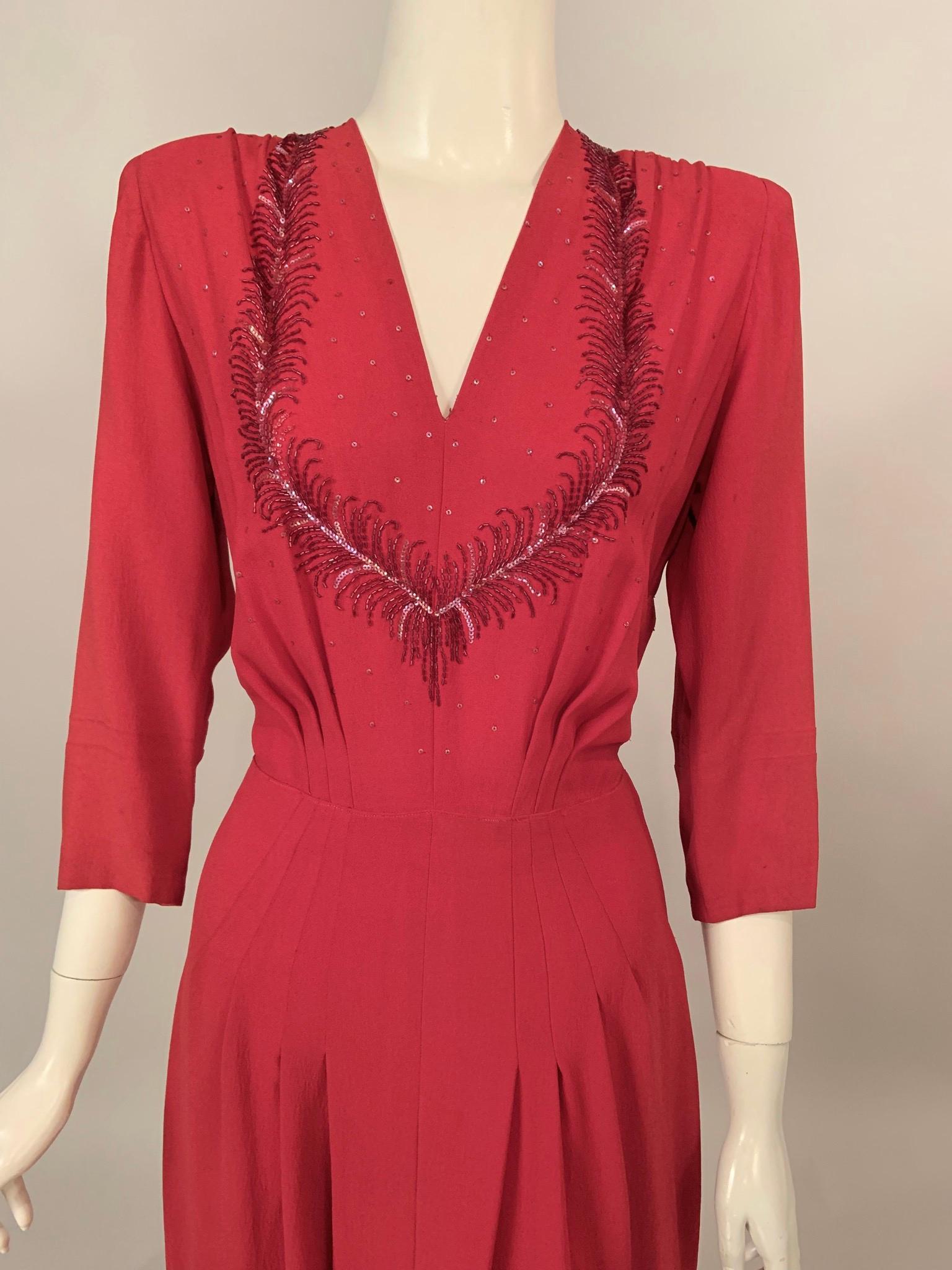 This 1940's dress is a beautiful shade of red embellished with darker red bugle beads and miniature opalescent sequins surrounding the low neckline. The dress has loose pleats on the bodice above stitched down pleats over the hips adding a sleek