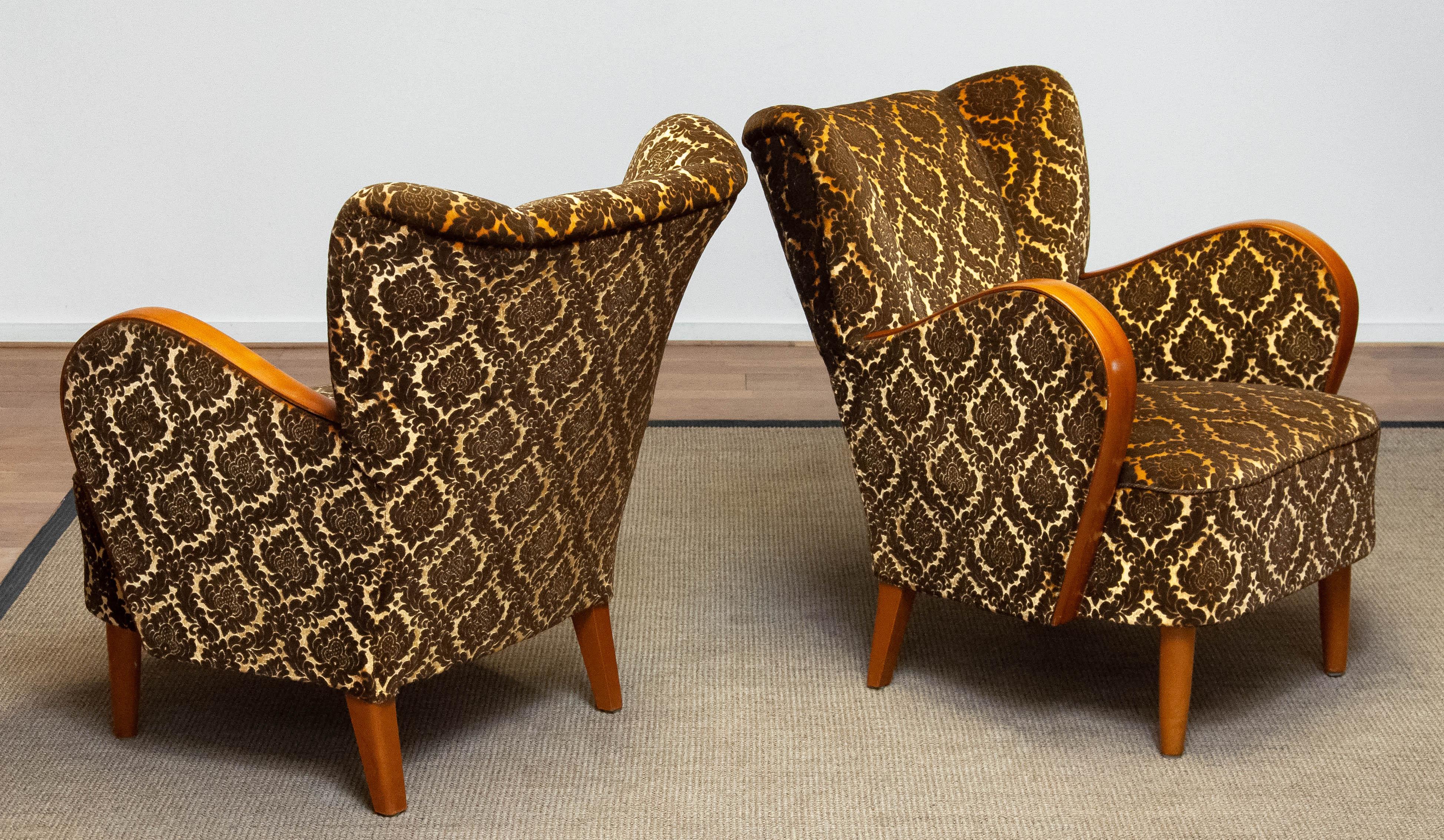 Preciosa y completa pareja de sillones escandinavos de estilo Fritz Hansen fabricados en Dinamarca.
Ambos sillones de salón / club están tapizados con tela de terciopelo jacquard bicolor dorado y marrón. Los reposabrazos de madera curvada son de