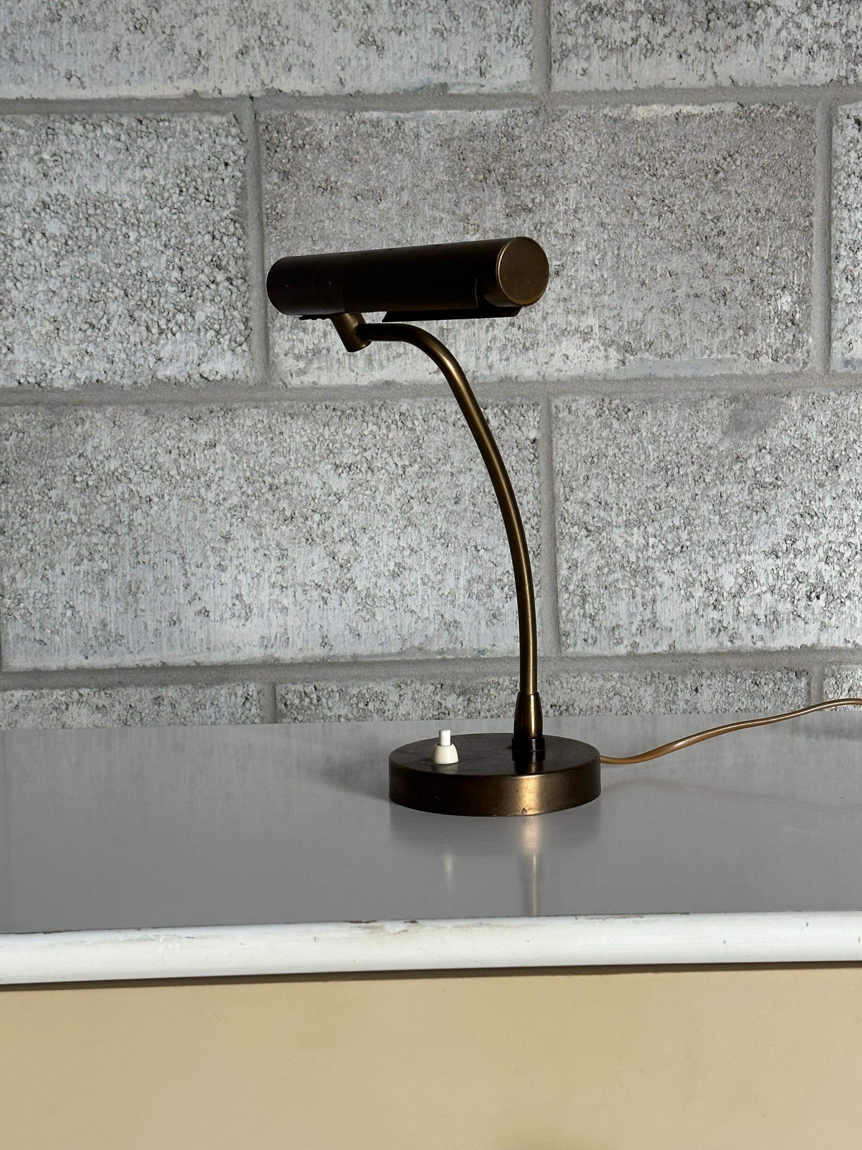 A wonderful brass lamp produced by Asea, very much in the style of a desk or banker lamp. Features a weighted brass base with push on/off button. An elongated curved stem continues up and to the side until reach the shade. Lamp has some degrees of
